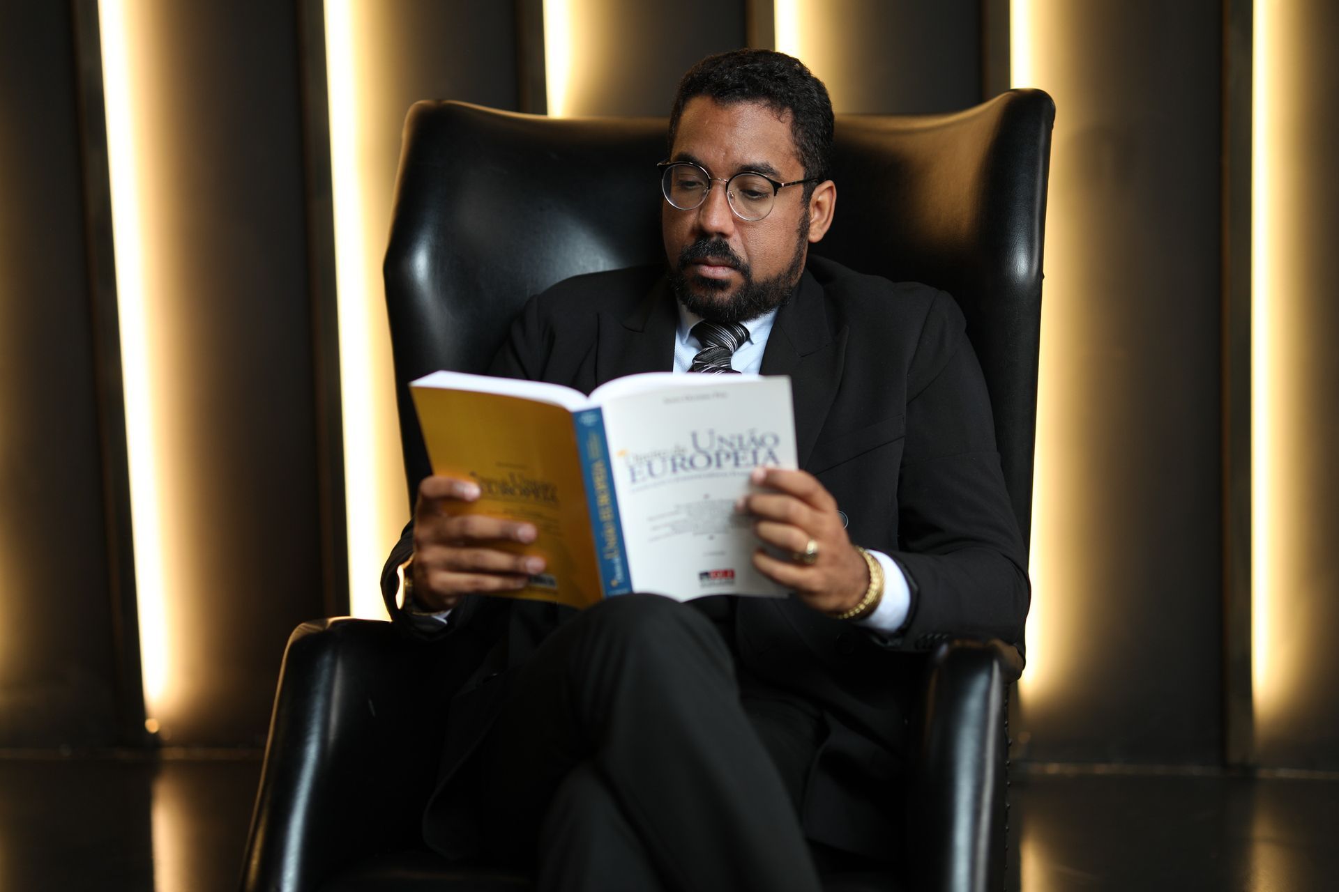 Homem de terno lê um livro sentado em uma poltrona de couro preta. Luz quente ao fundo.