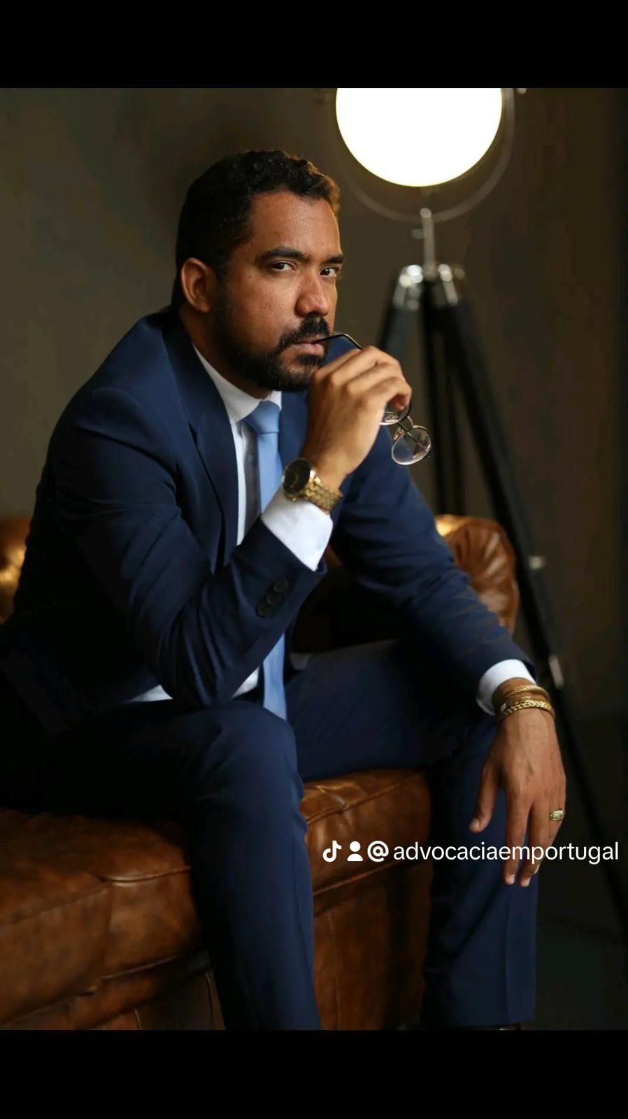 Homem de terno azul, segurando um copo, está sentado em um sofá de couro, com um olhar pensativo perto de um abajur.