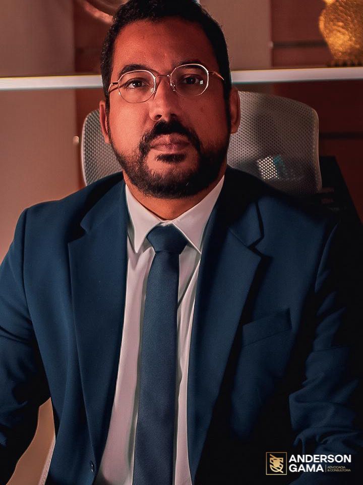 Homem de terno azul e óculos sentado, olhando para a frente. Ambiente de escritório.