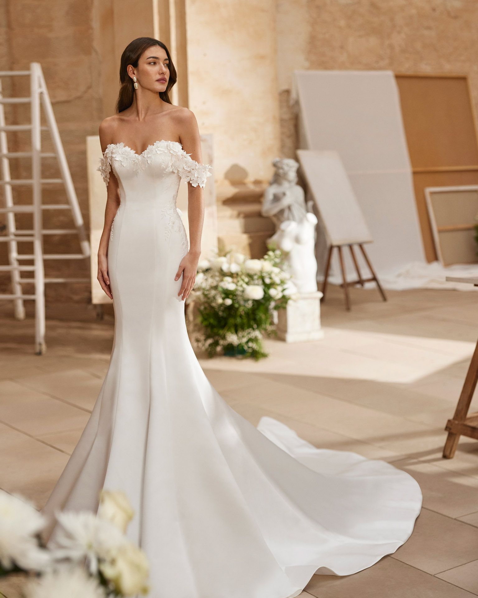Donna in abito da sposa bianco a sirena con spalle scoperte, corpetto floreale, strascico, in piedi in una stanza piena di luce.
