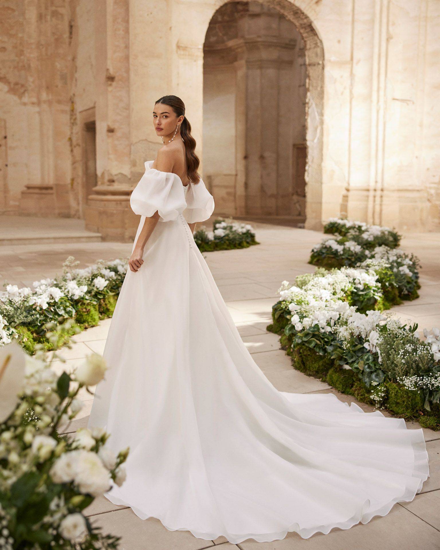 Donna in abito da sposa bianco con spalle scoperte e lungo strascico, in piedi in uno spazio luminoso e arcuato.