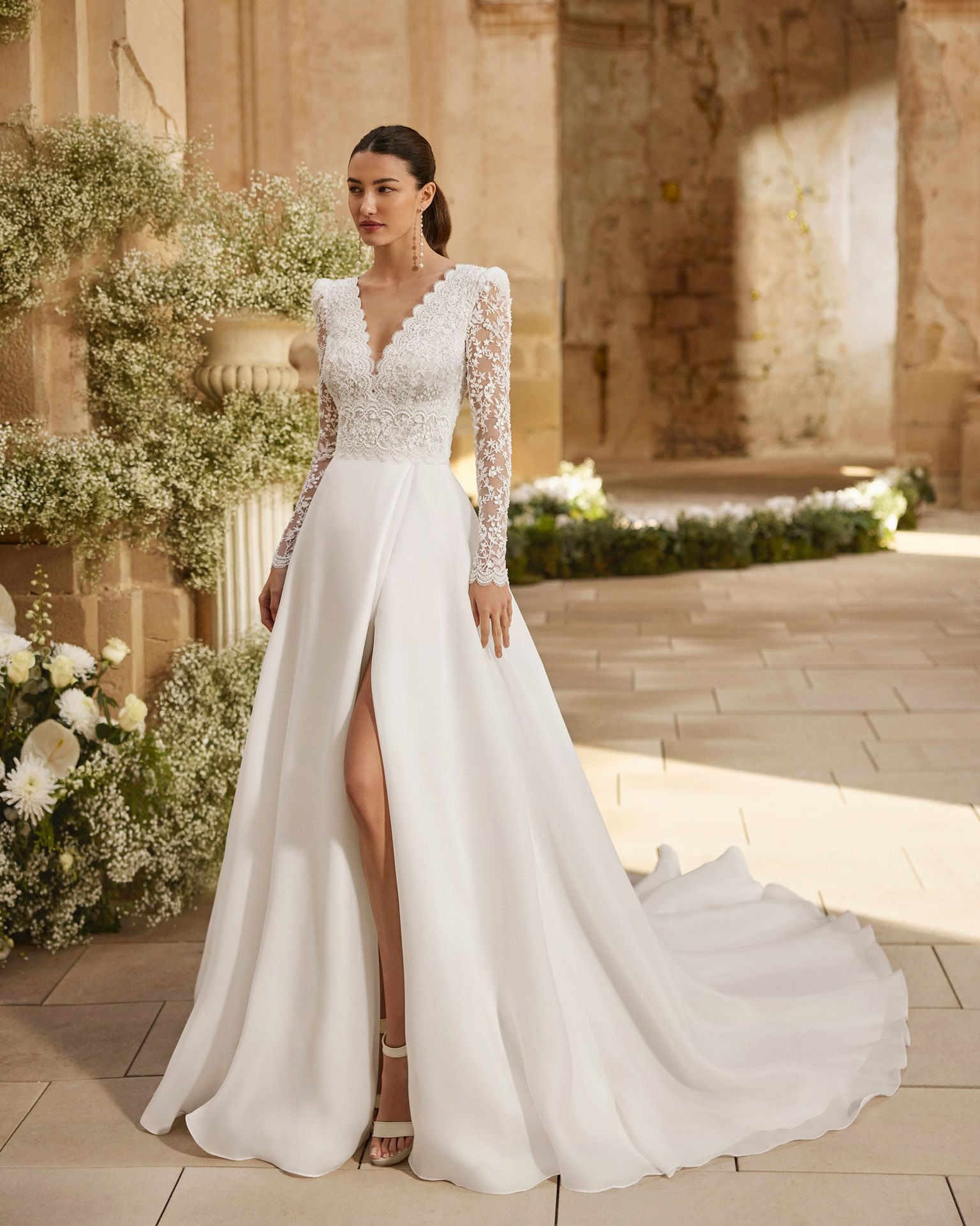 Donna in abito da sposa bianco con spacco profondo e maniche lunghe, in piedi all'esterno.