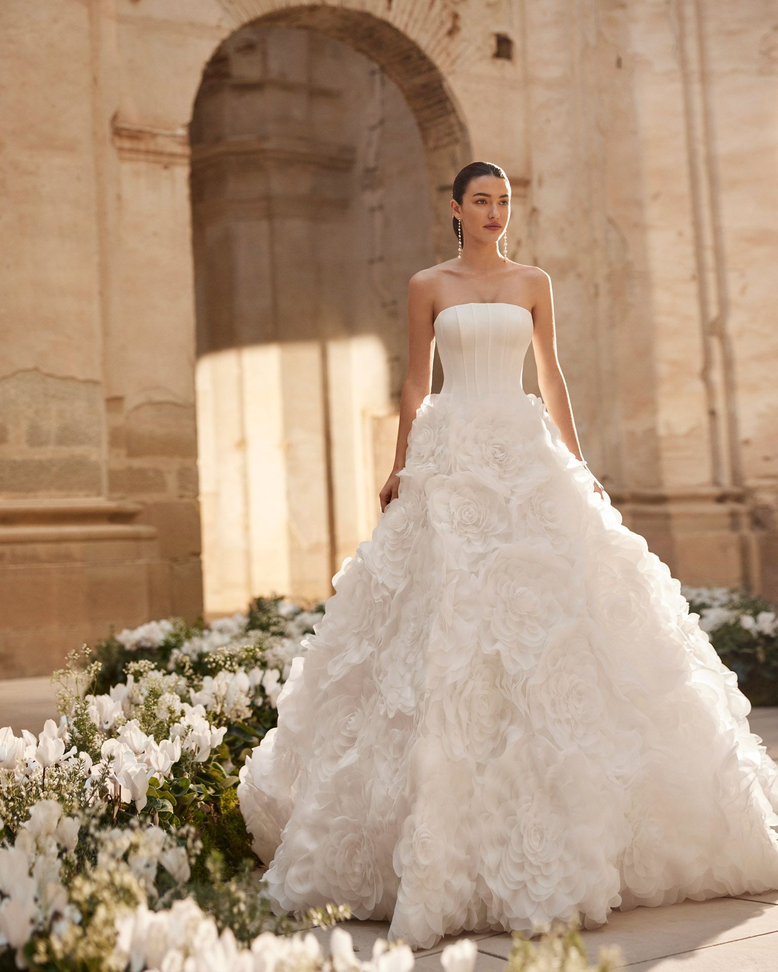 Donna in abito da sposa bianco senza spalline in posa vicino a fiori e architettura in pietra.