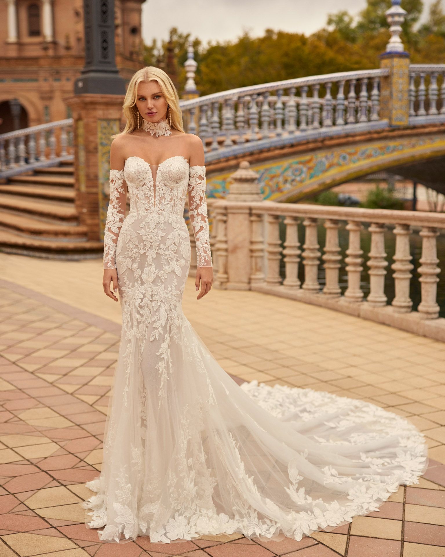Donna con abito da sposa in pizzo bianco con lungo strascico, in piedi su un sentiero di mattoni, con un ponte sullo sfondo.