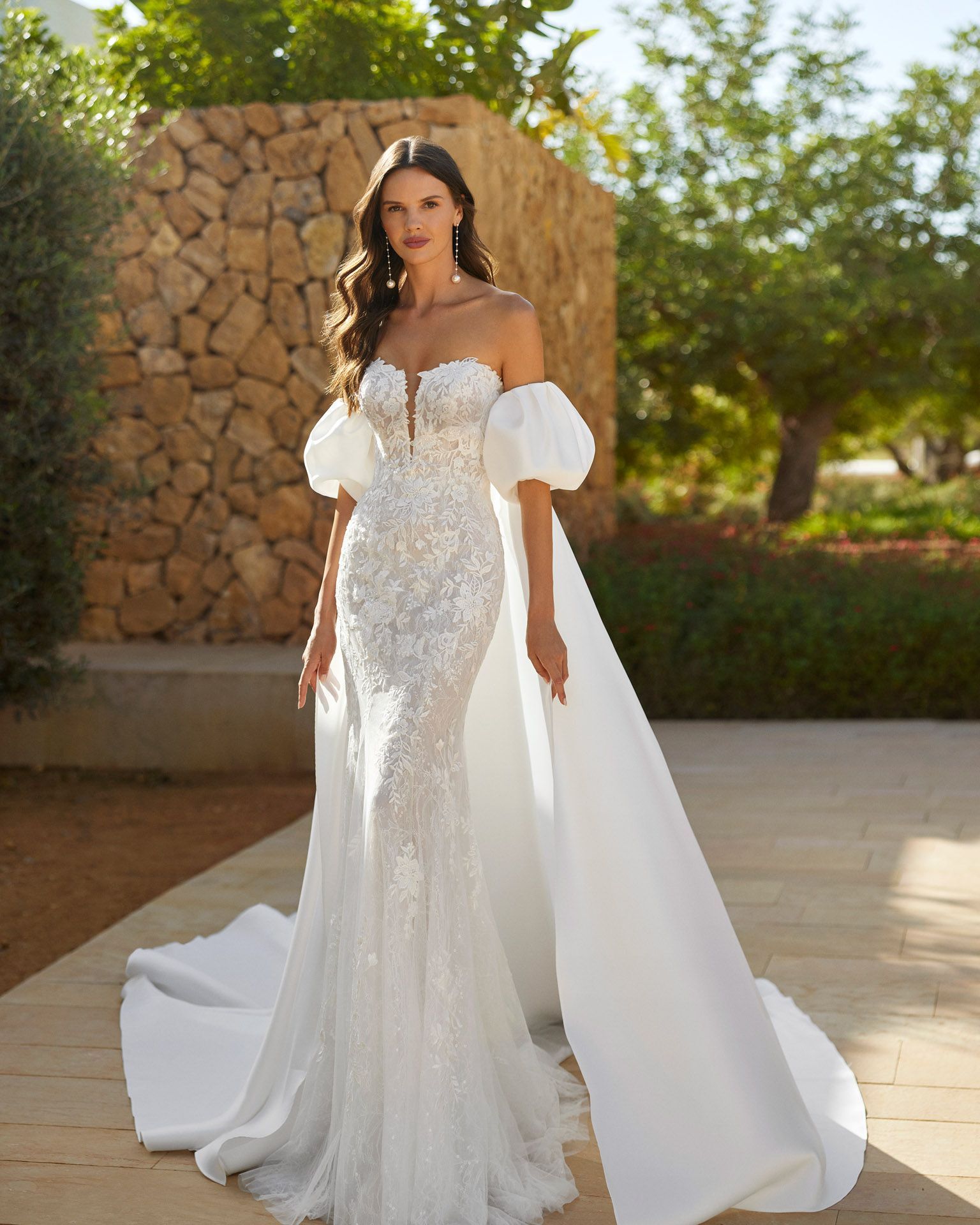 Donna in abito da sposa bianco con perline, strascico e maniche a sbuffo, in posa all'aperto.