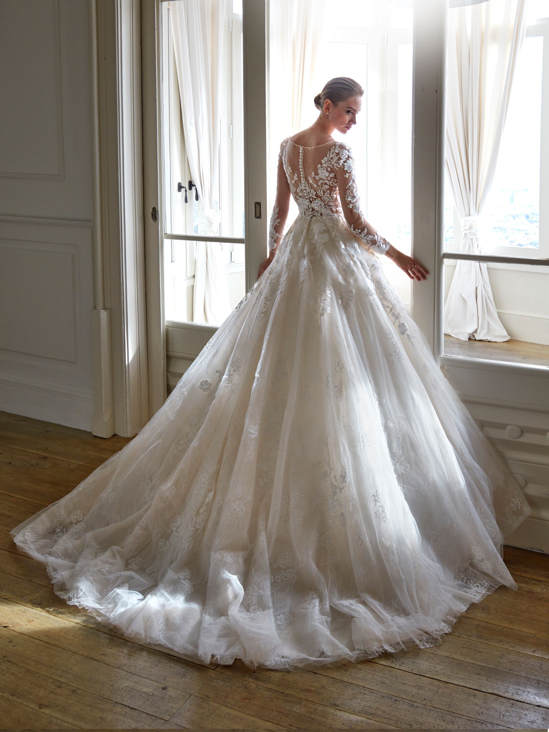Una donna in abito da sposa bianco con lungo strascico è in piedi vicino a una finestra.