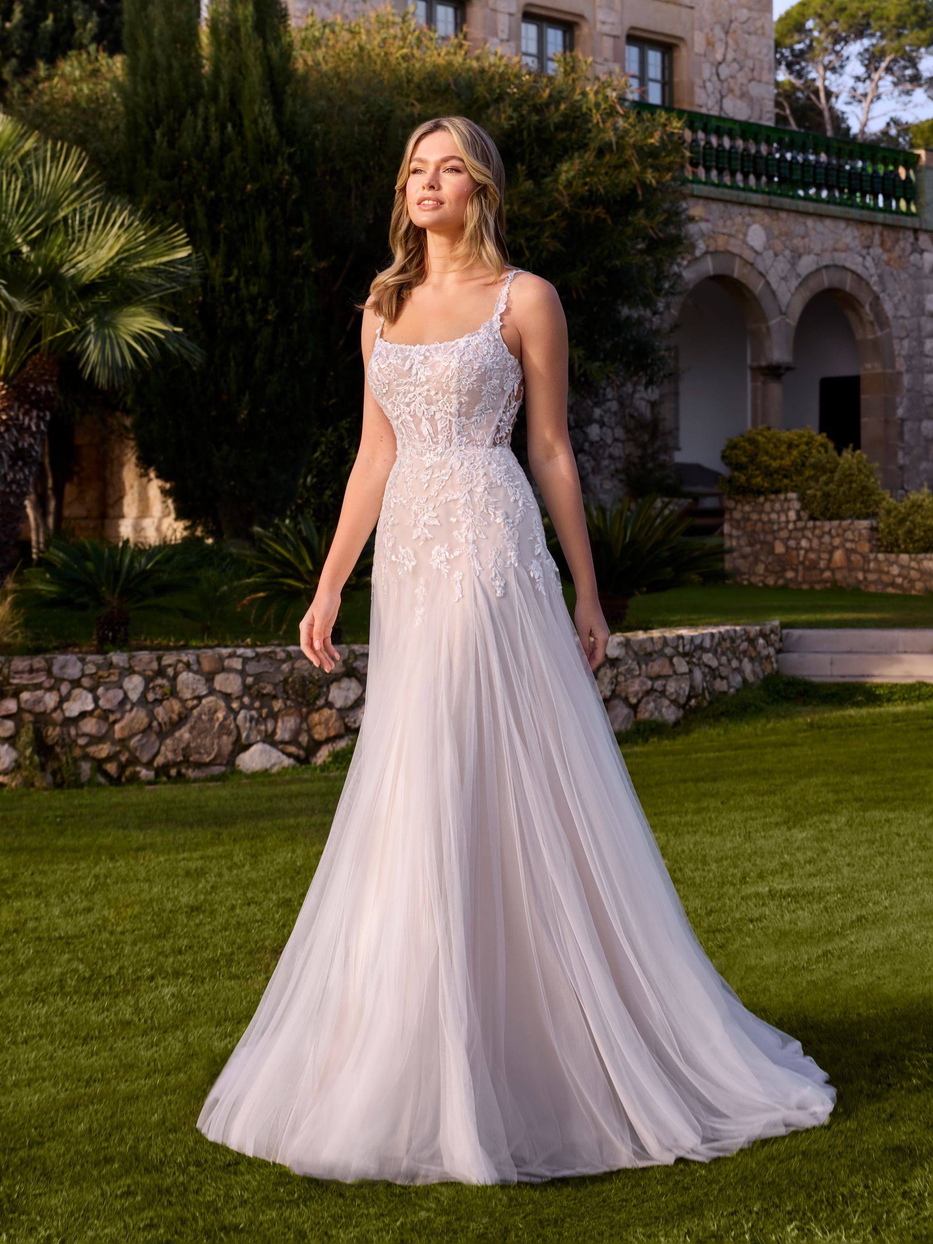 Donna in abito da sposa bianco davanti a un edificio in pietra con erba verde.