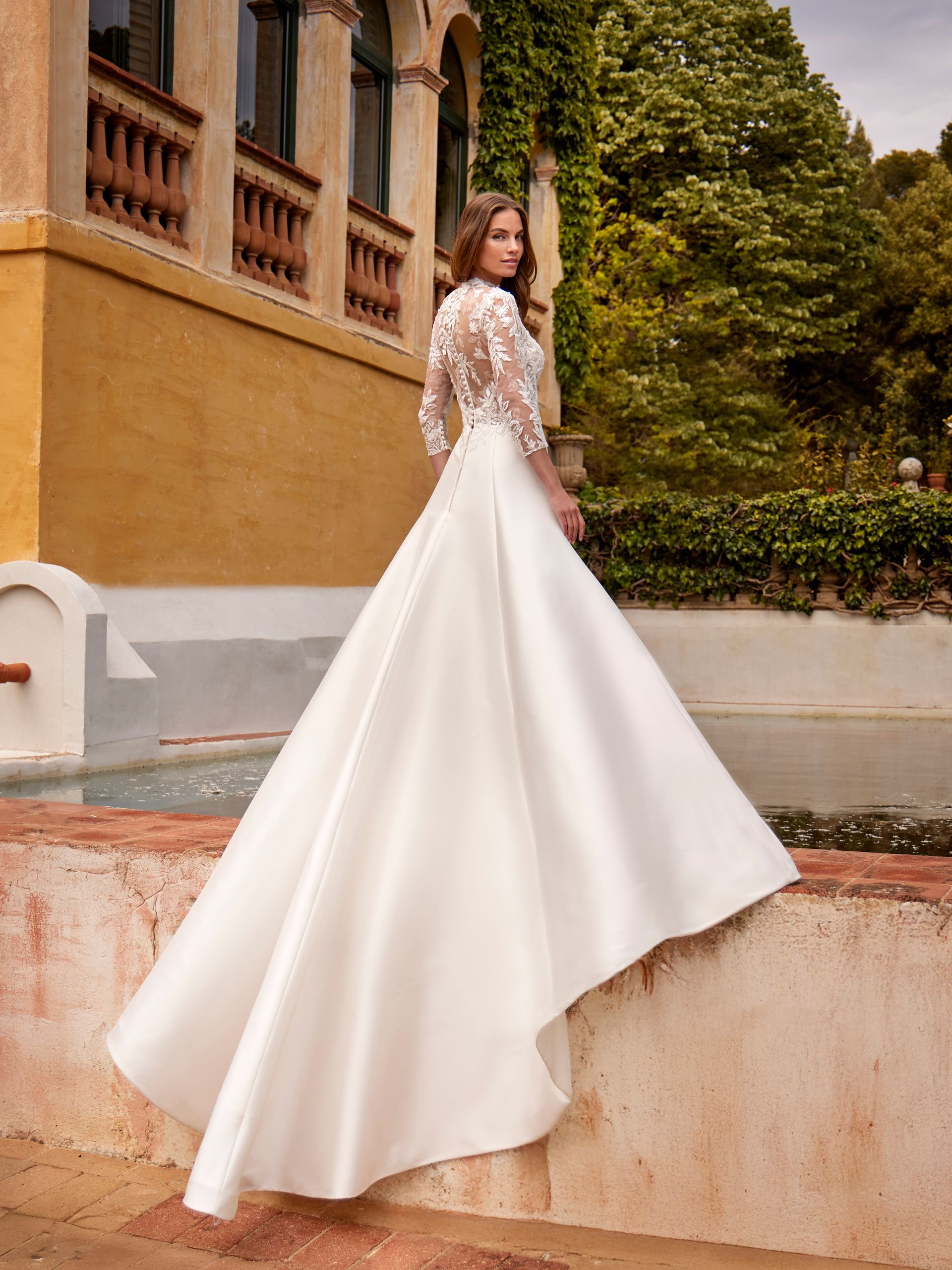 Sposa in abito da sposa in raso bianco con maniche in pizzo, in piedi all'esterno.