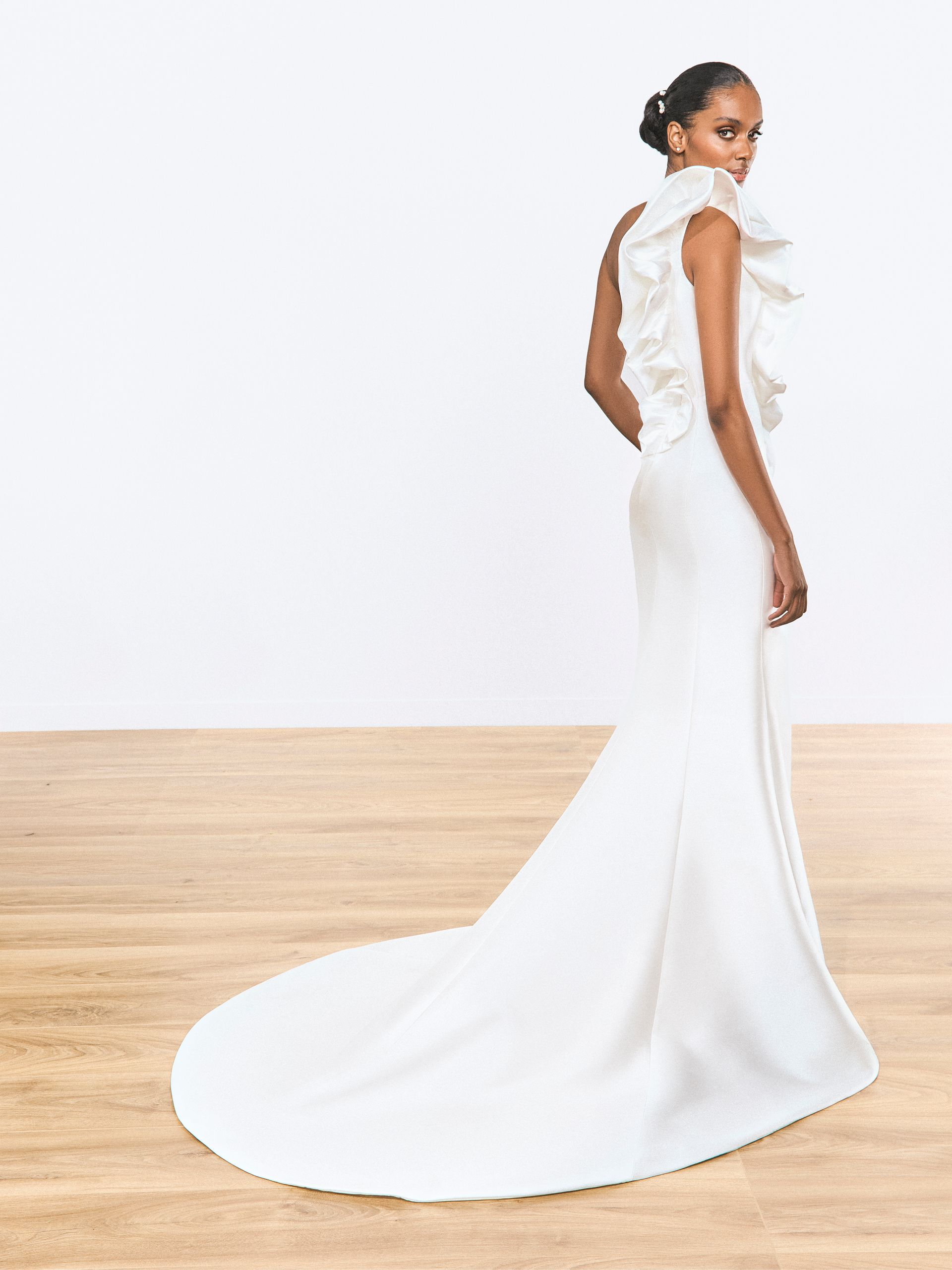 Donna in abito da sposa bianco con spalle arricciate e lungo strascico, in piedi su un pavimento di legno.