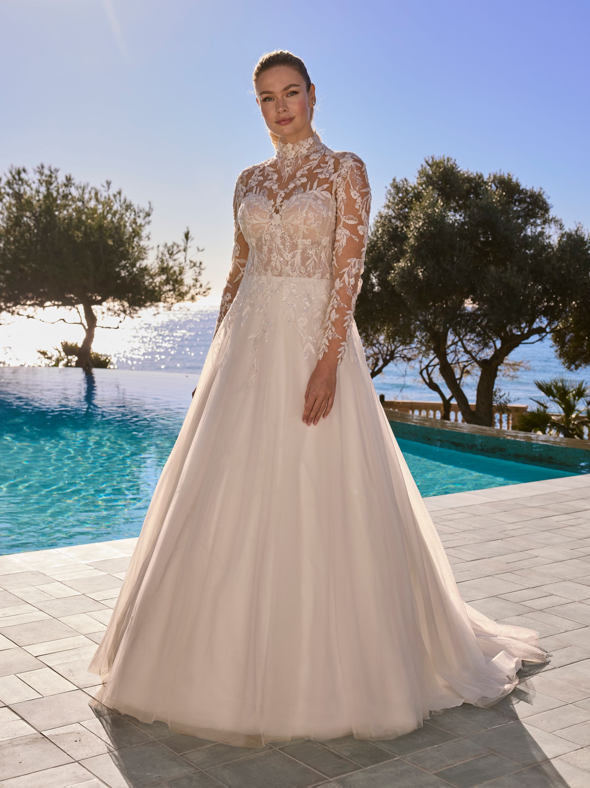 Donna in abito da sposa, in piedi vicino a una piscina. L'abito è bianco con maniche in pizzo. Scenario esterno soleggiato.