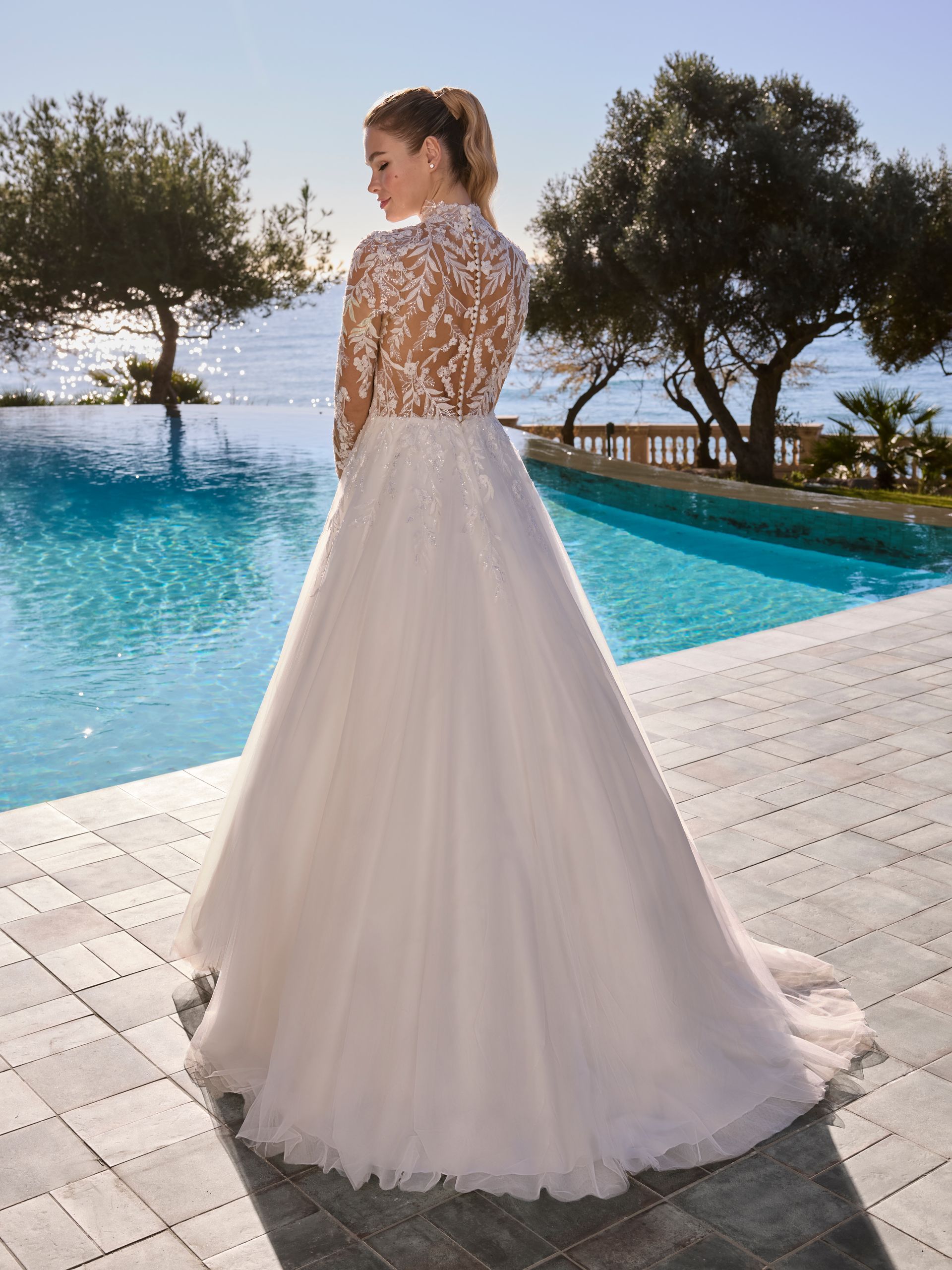 Donna in abito da sposa bianco, corpetto di pizzo, in piedi vicino alla piscina con vista sul mare.