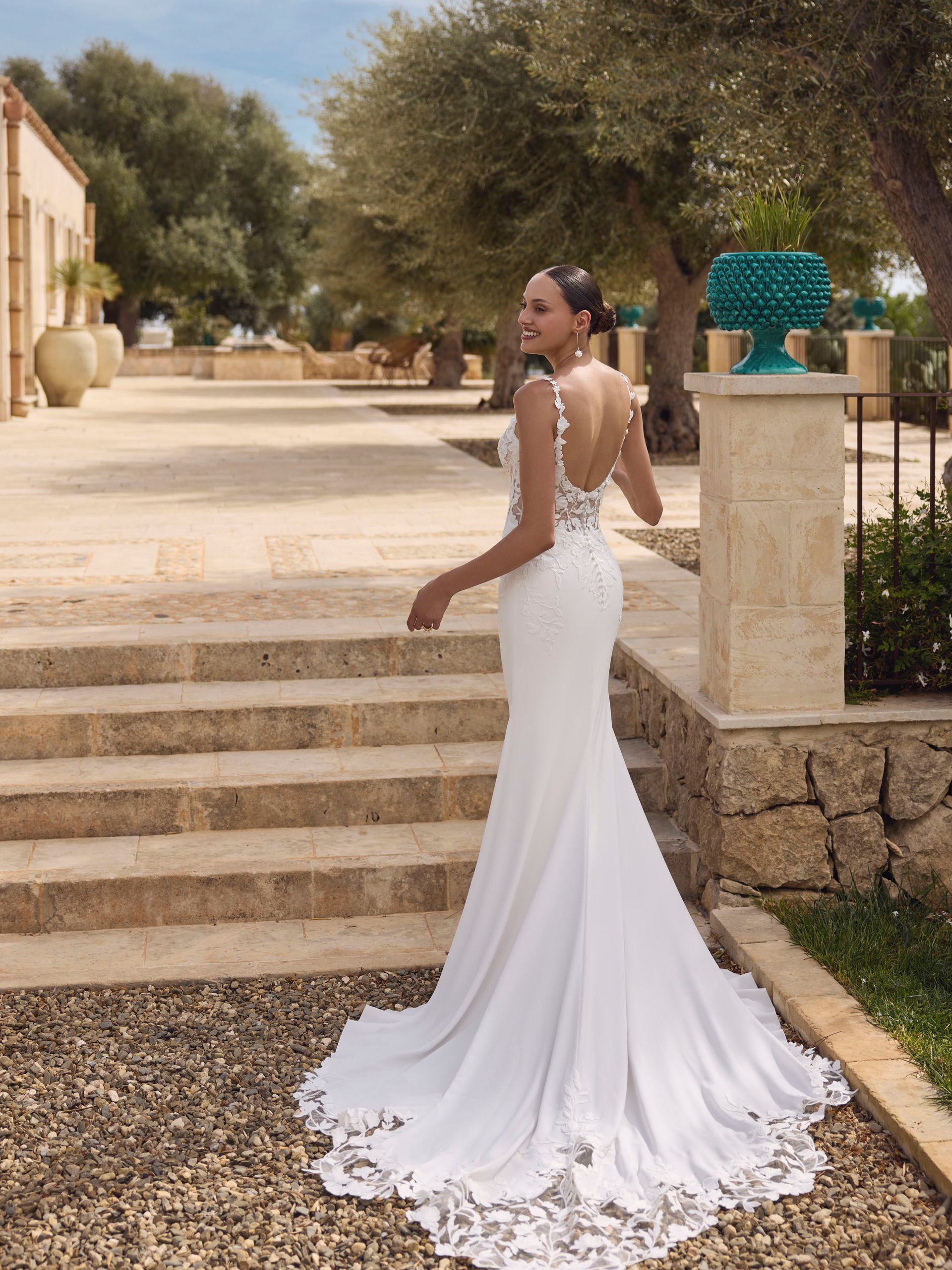 Una donna in abito da sposa bianco, sorridente, posa sui gradini di pietra all'esterno; ambiente soleggiato e mediterraneo.