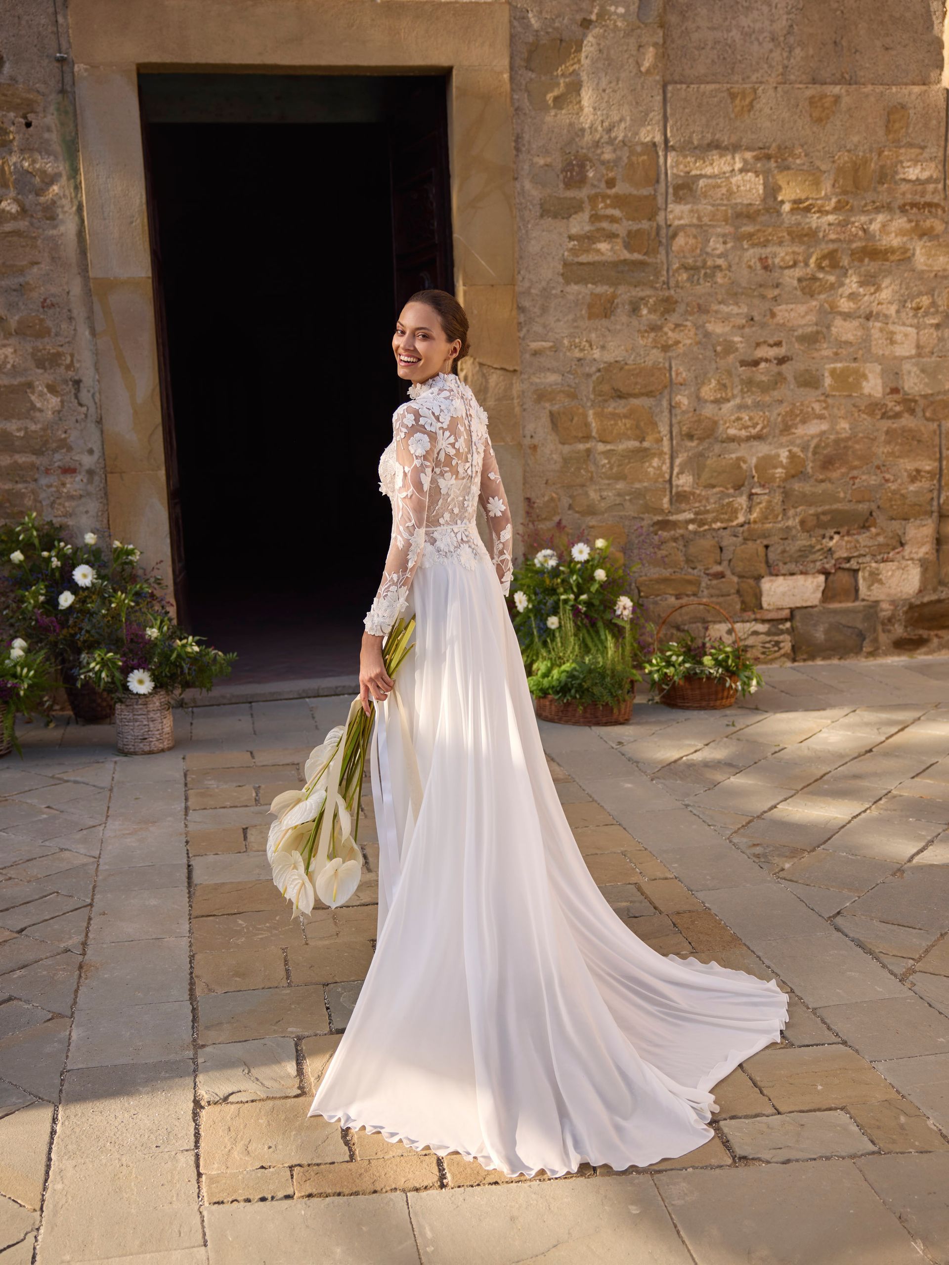 La sposa, con un abito di pizzo, sorride mentre si allontana da un edificio in pietra, tenendo in mano dei fiori.