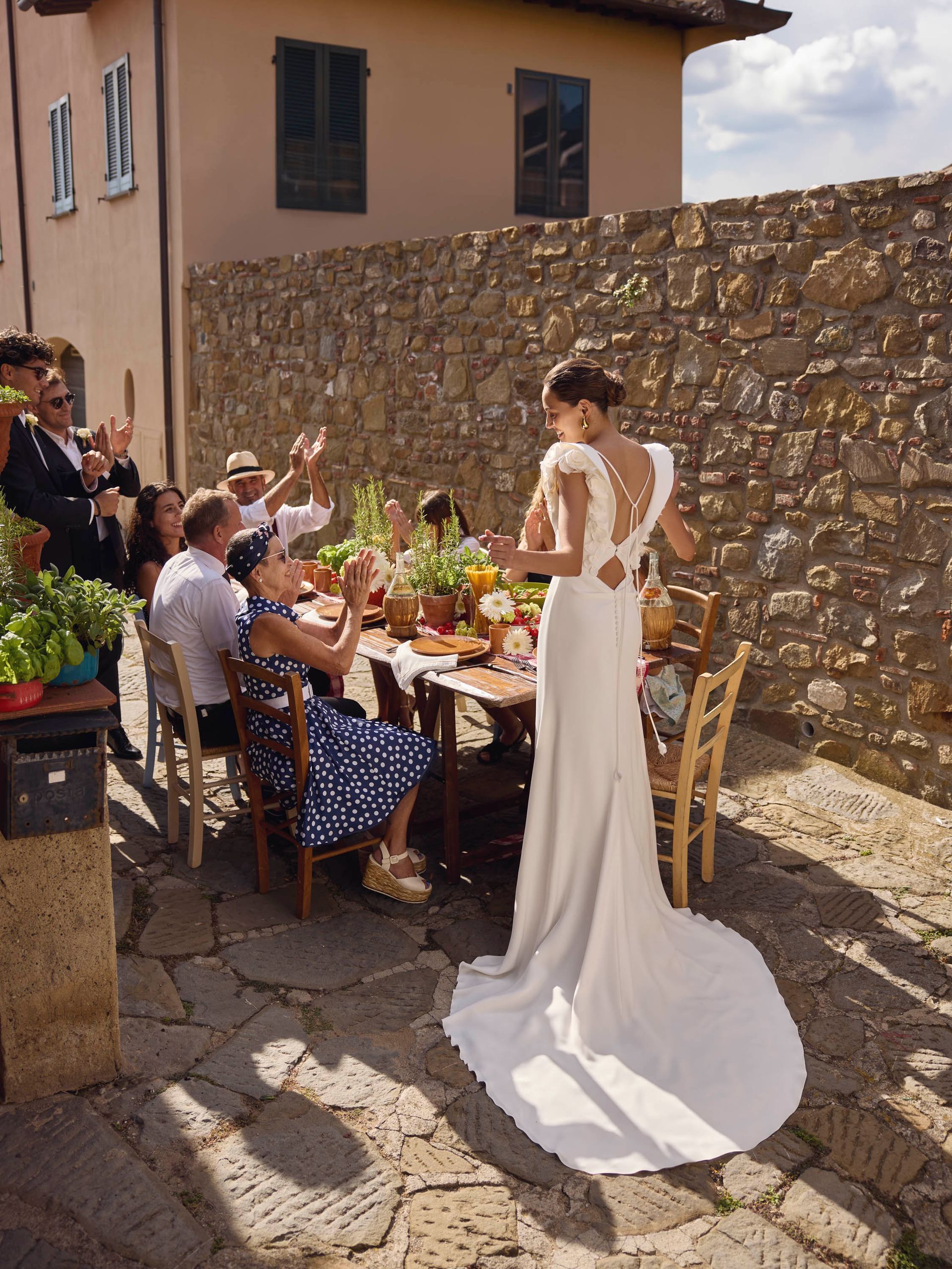 Sposa in abito bianco con strascico, accoglie gli ospiti seduti al tavolo da pranzo all'aperto. Cortile in pietra.