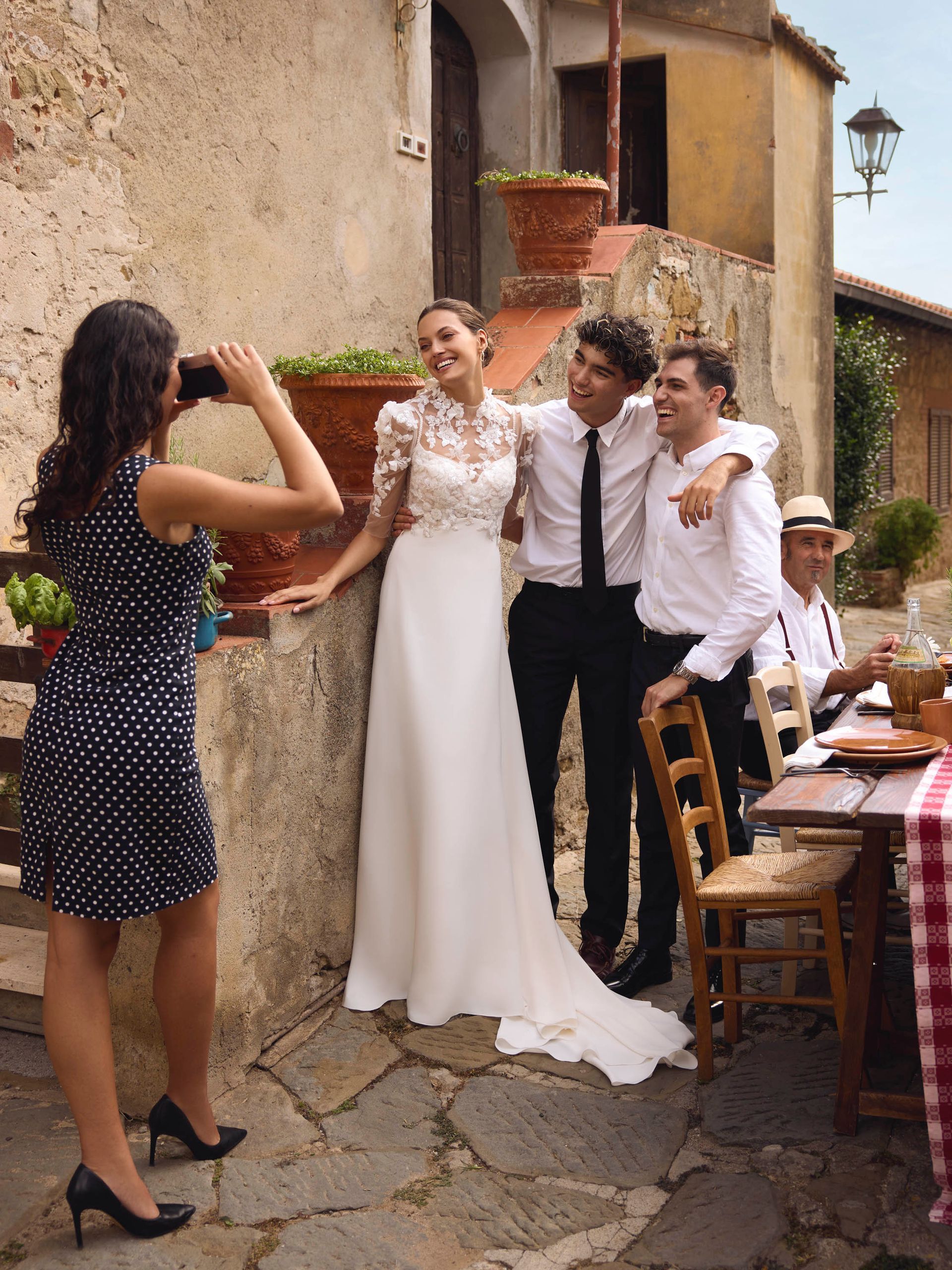 La sposa posa con due uomini mentre una donna scatta una foto. L'ambientazione è un bar all'aperto su una strada lastricata.