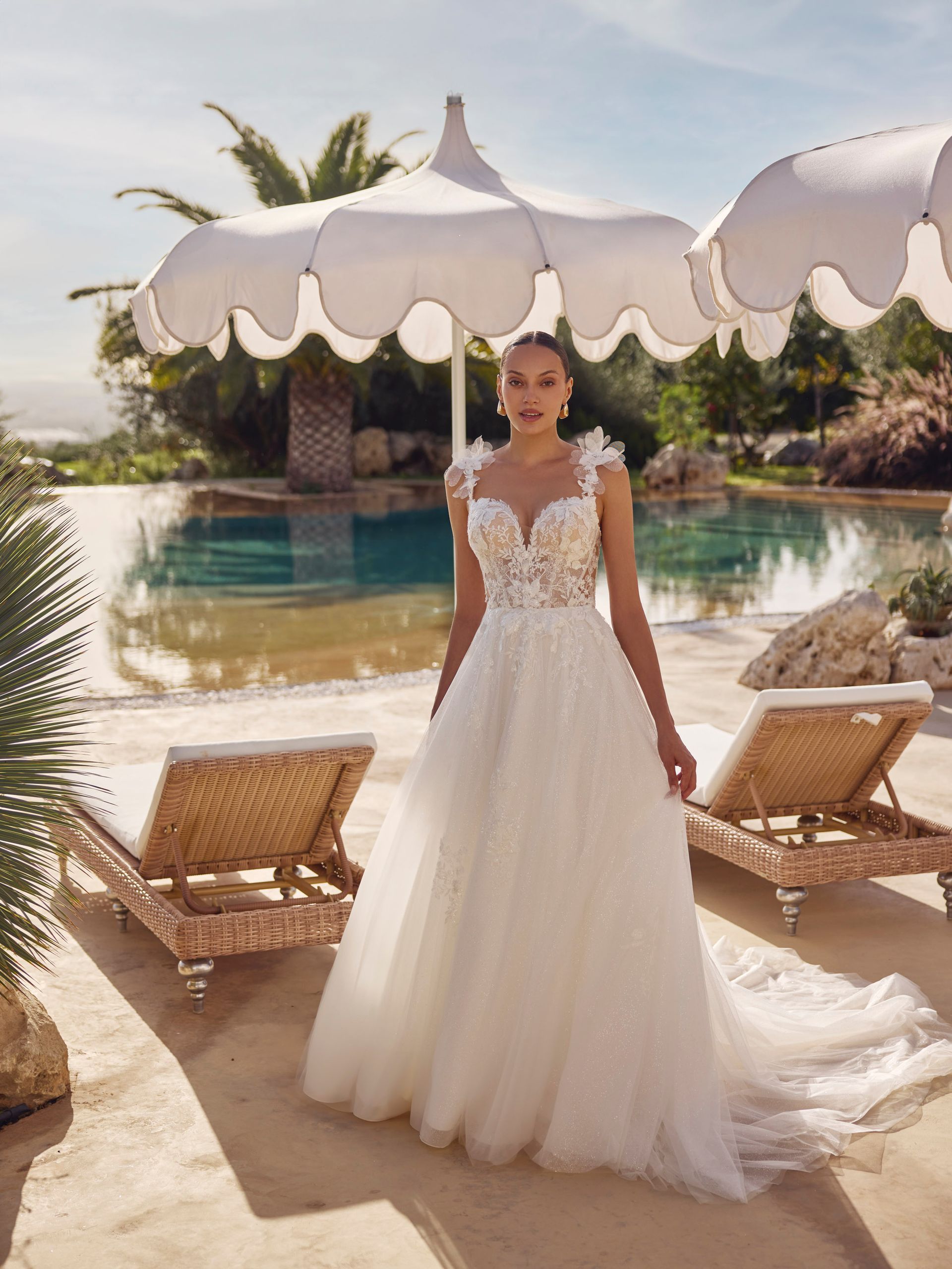 Una donna in abito da sposa bianco è in piedi vicino a una piscina, sotto un ombrellone, e sorride.
