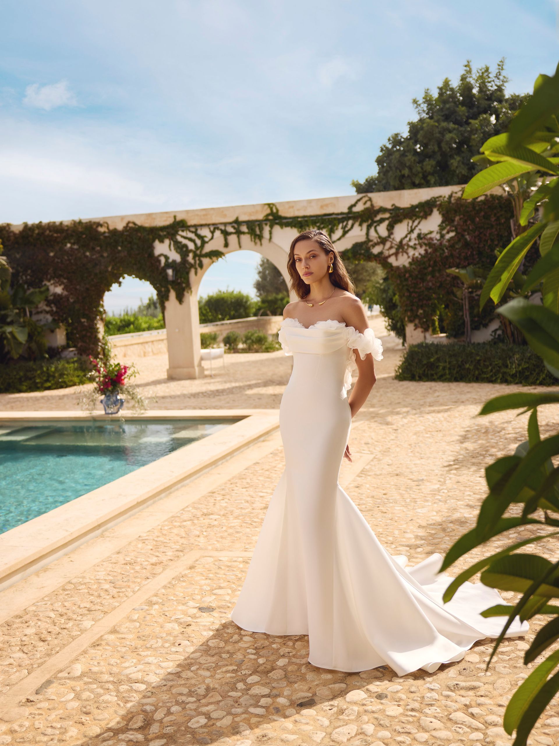 Una donna con un abito da sposa bianco a spalle scoperte posa vicino a una piscina, in un giardino.
