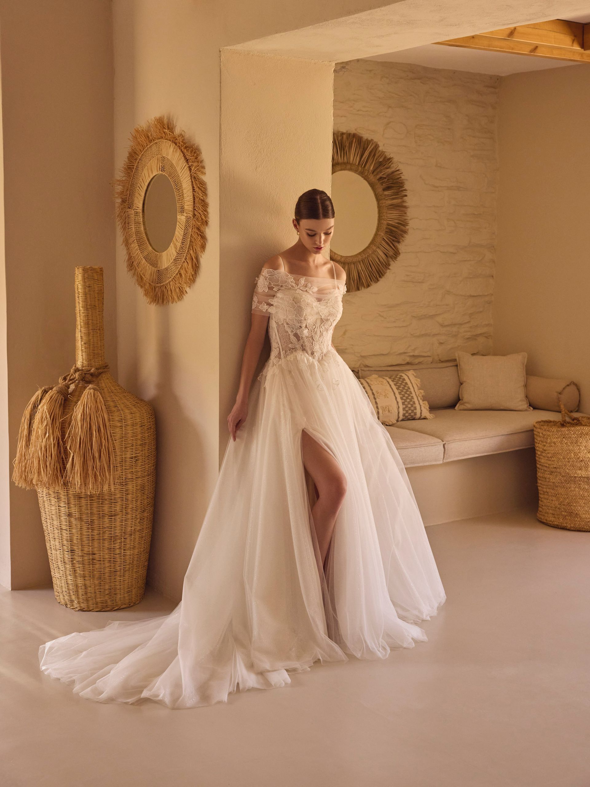 Donna in abito da sposa bianco con spacco profondo, in piedi in una stanza beige con decorazioni intrecciate.