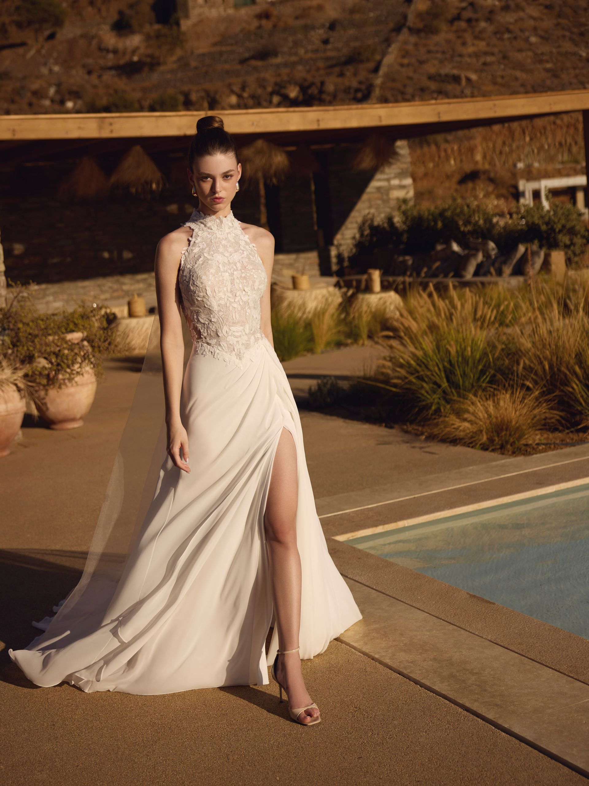 Donna in abito da sposa bianco, con spacco profondo, in piedi a bordo piscina.