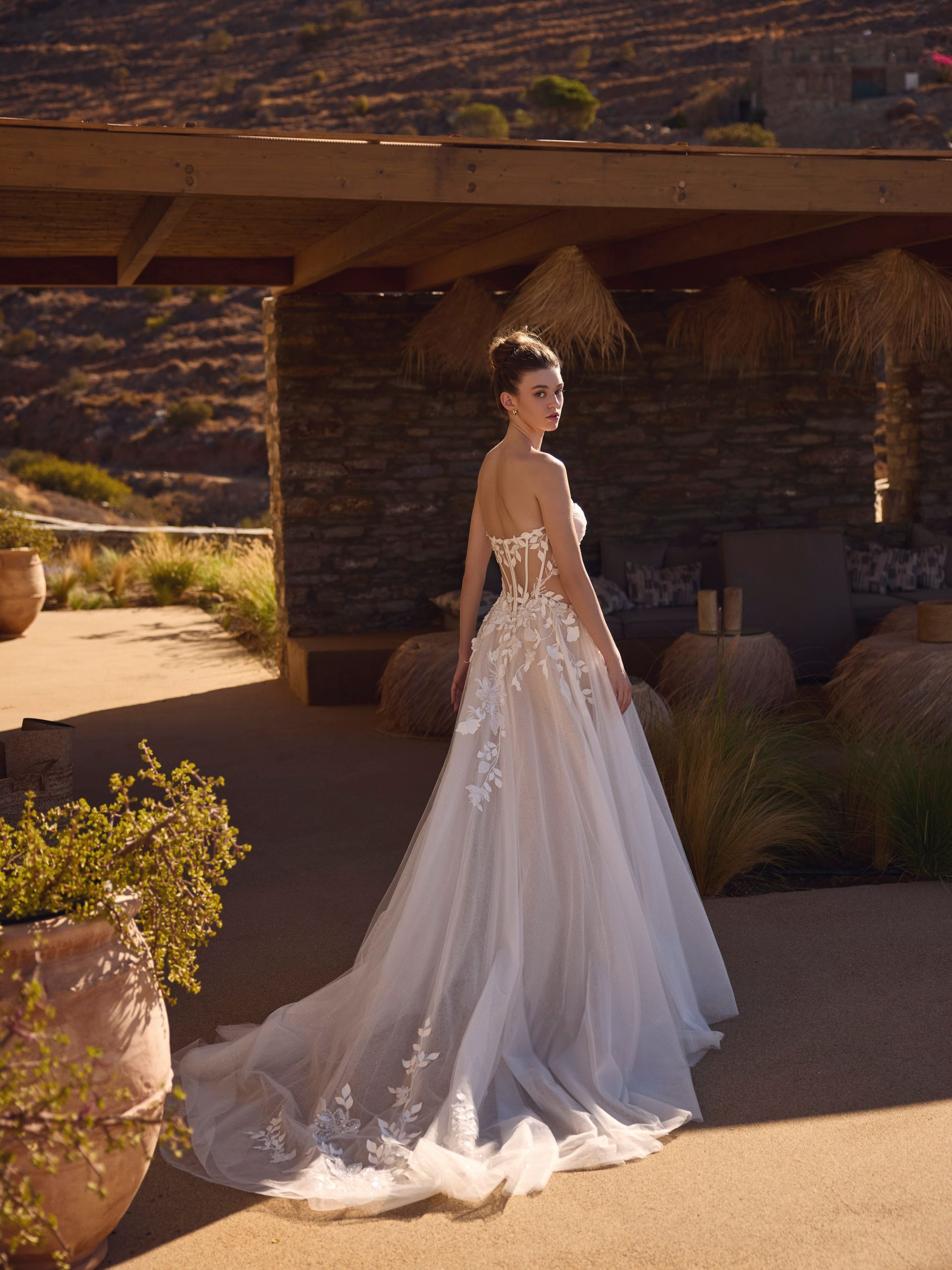 Donna in abito da sposa, che guarda indietro, in piedi all'esterno sotto un pergolato con una collina sullo sfondo.
