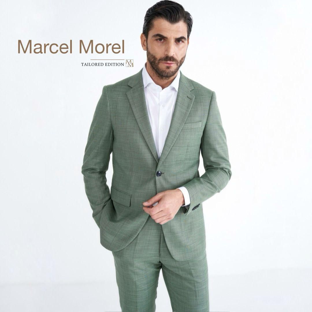 Uomo in abito verde, camicia bianca, mani in tasca, in posa su sfondo bianco.