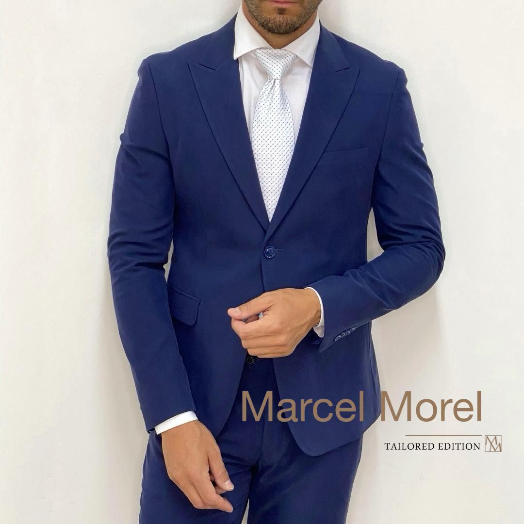 Uomo in abito blu navy, camicia bianca e cravatta fantasia, in un ambiente interno, con in mano la giacca del suo abito.