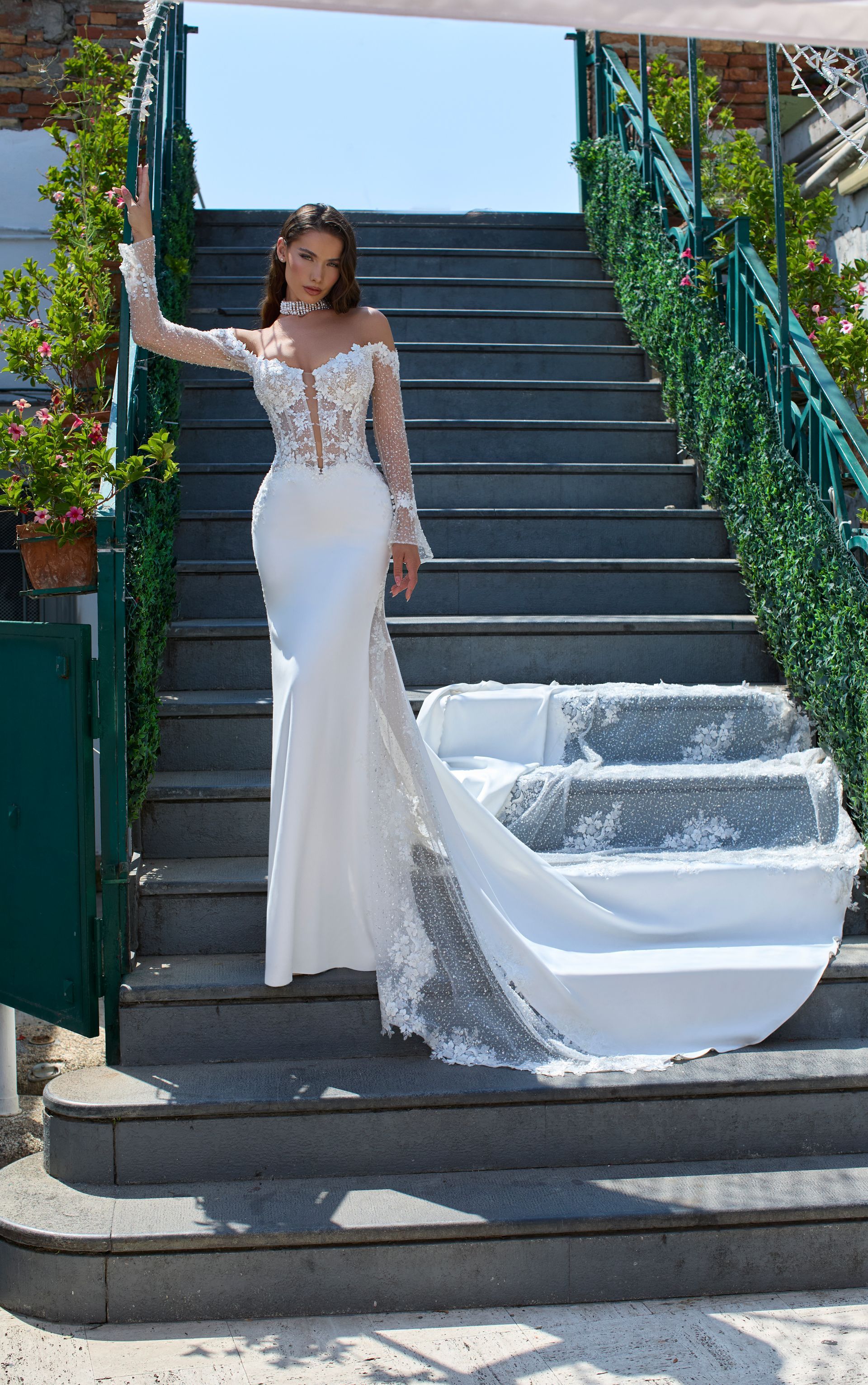 Sposa in abito da sposa bianco con spalle scoperte e lungo strascico, in posa sulle scale esterne.