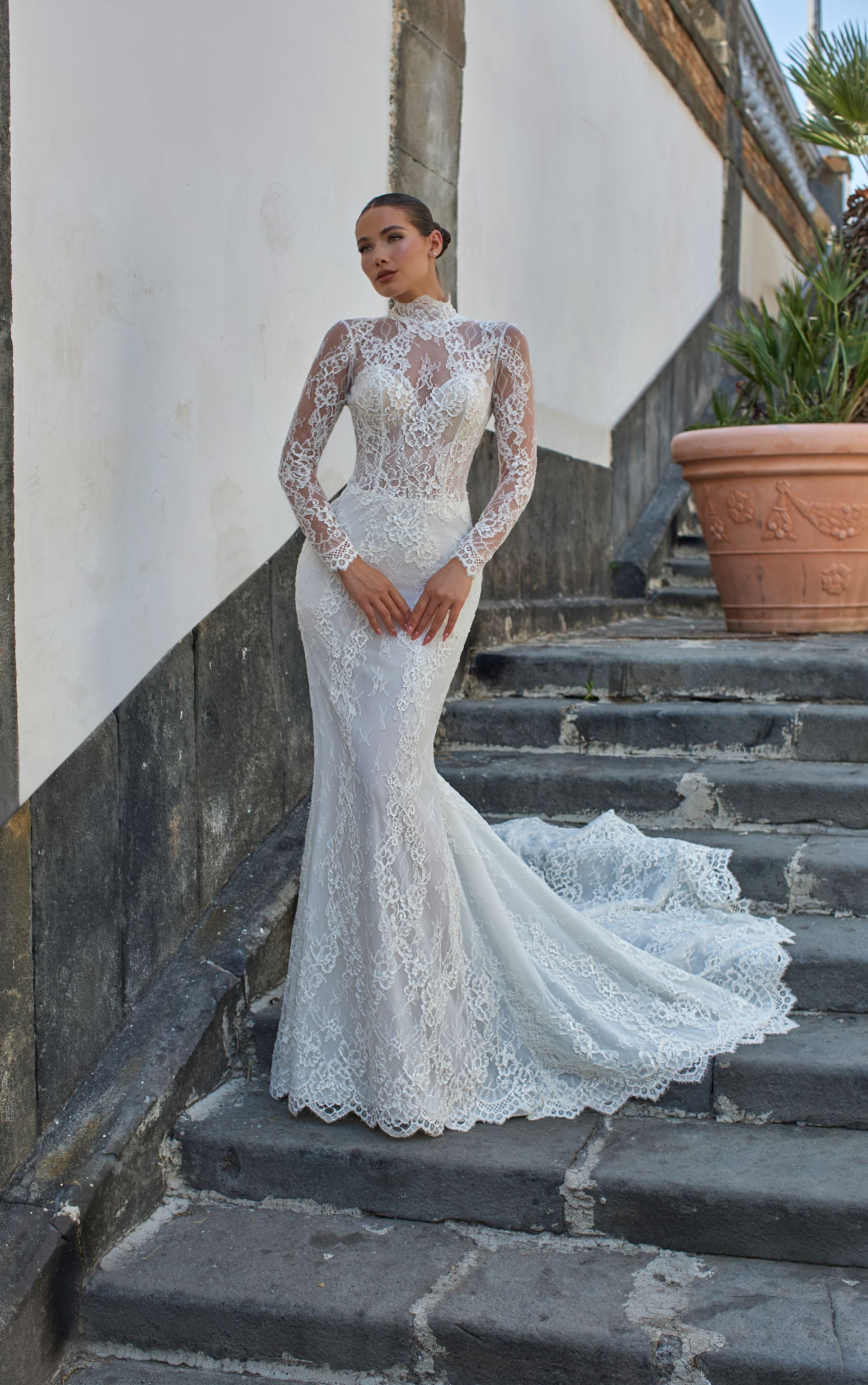 Donna in abito da sposa a sirena in pizzo bianco, con collo alto e lungo strascico, in piedi su gradini di pietra.