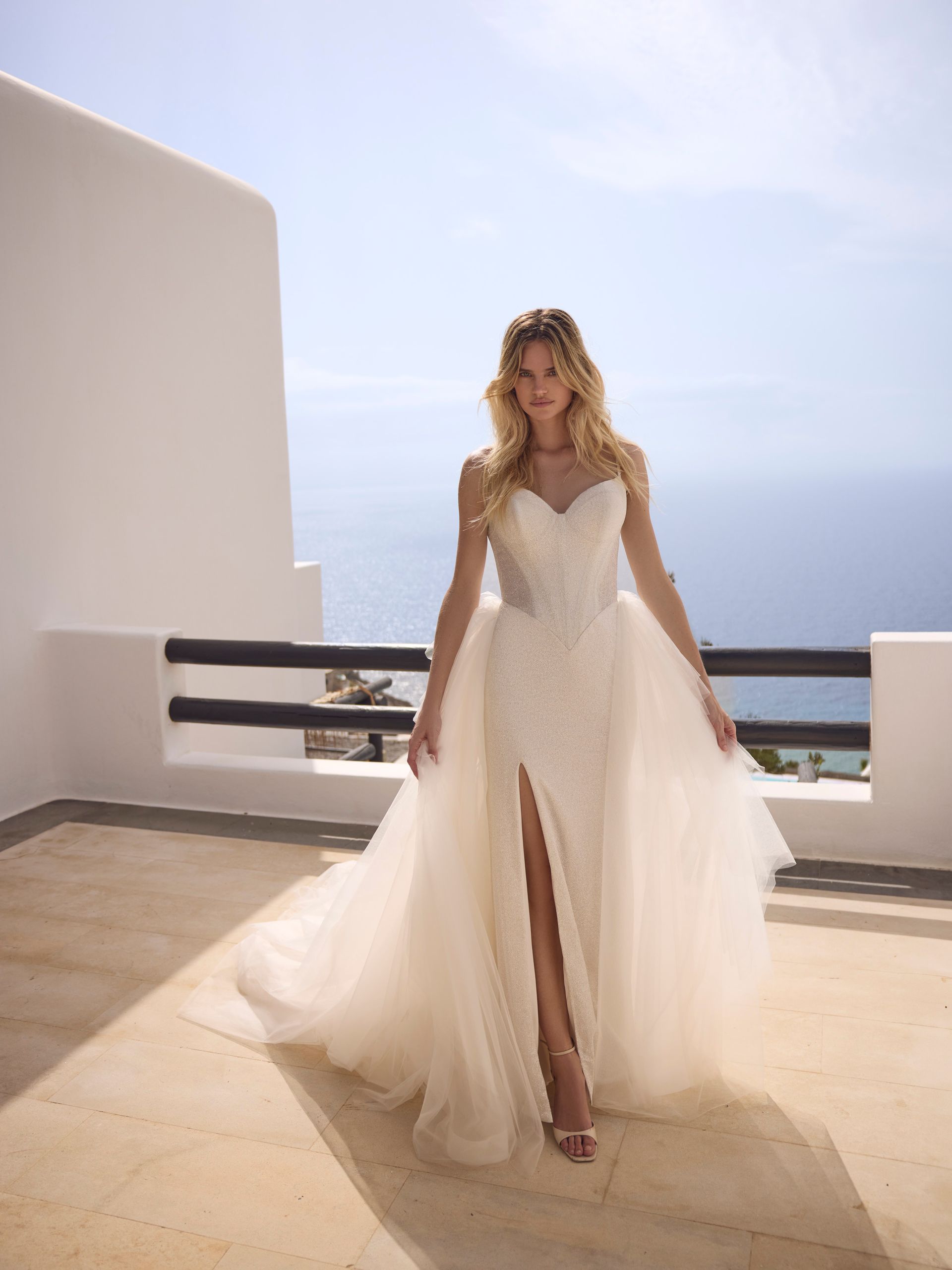 Una donna in abito da sposa con uno spacco profondo posa su un balcone bianco con vista sull'oceano.