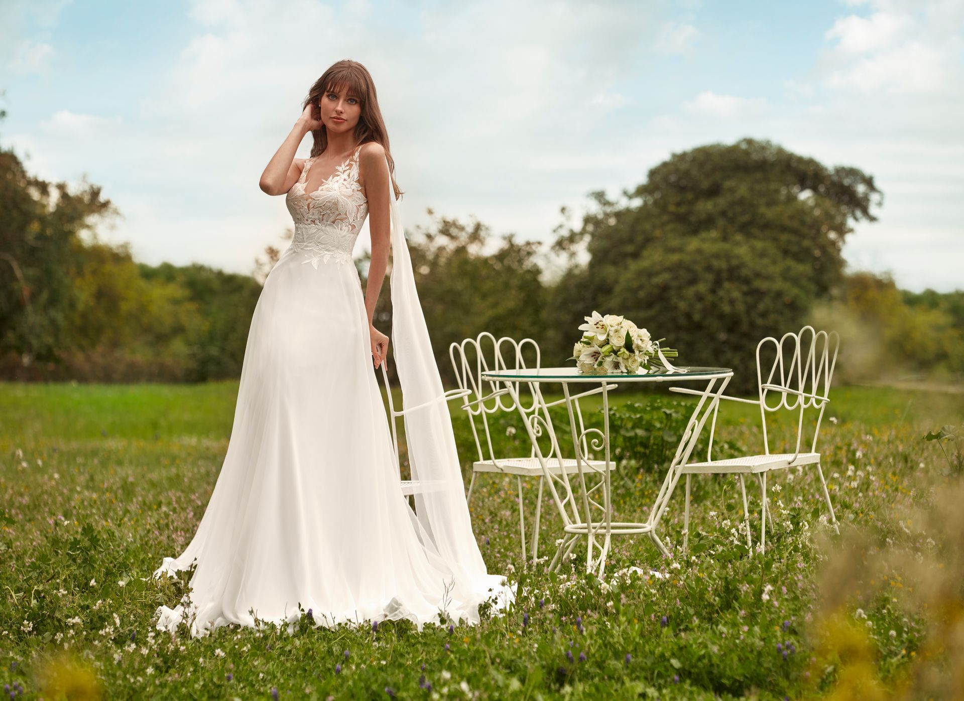 Donna in abito da sposa bianco in piedi in un campo accanto a un tavolino e delle sedie.