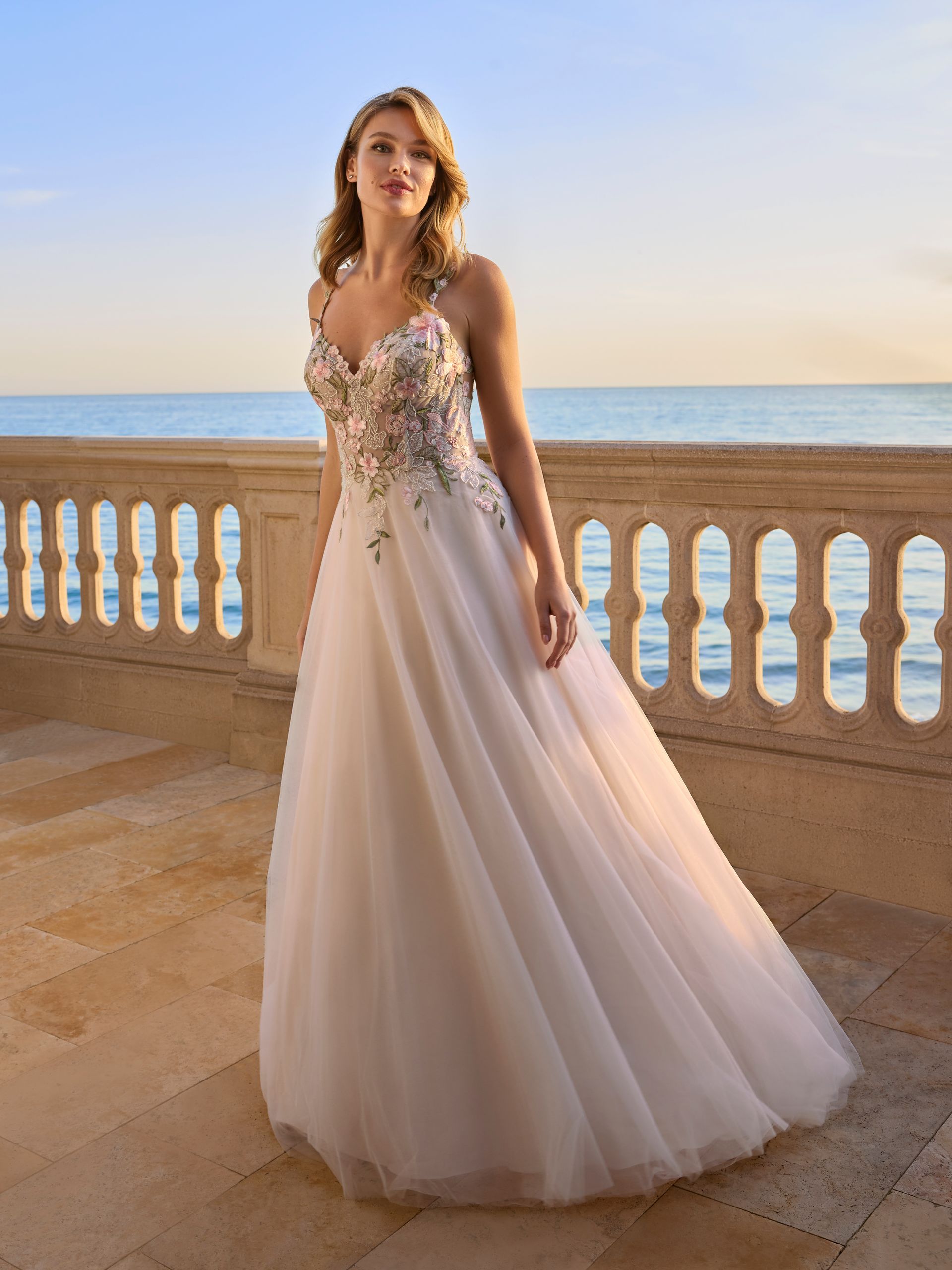Donna in abito da sposa sul balcone con vista sul mare; l'abito presenta dettagli floreali e colori tenui.