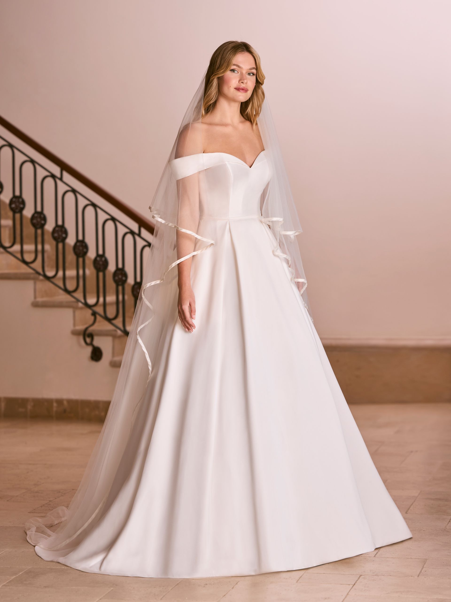 Sposa con abito da sposa in raso bianco con spalle scoperte e velo, in piedi su un pavimento di marmo vicino a una scalinata.