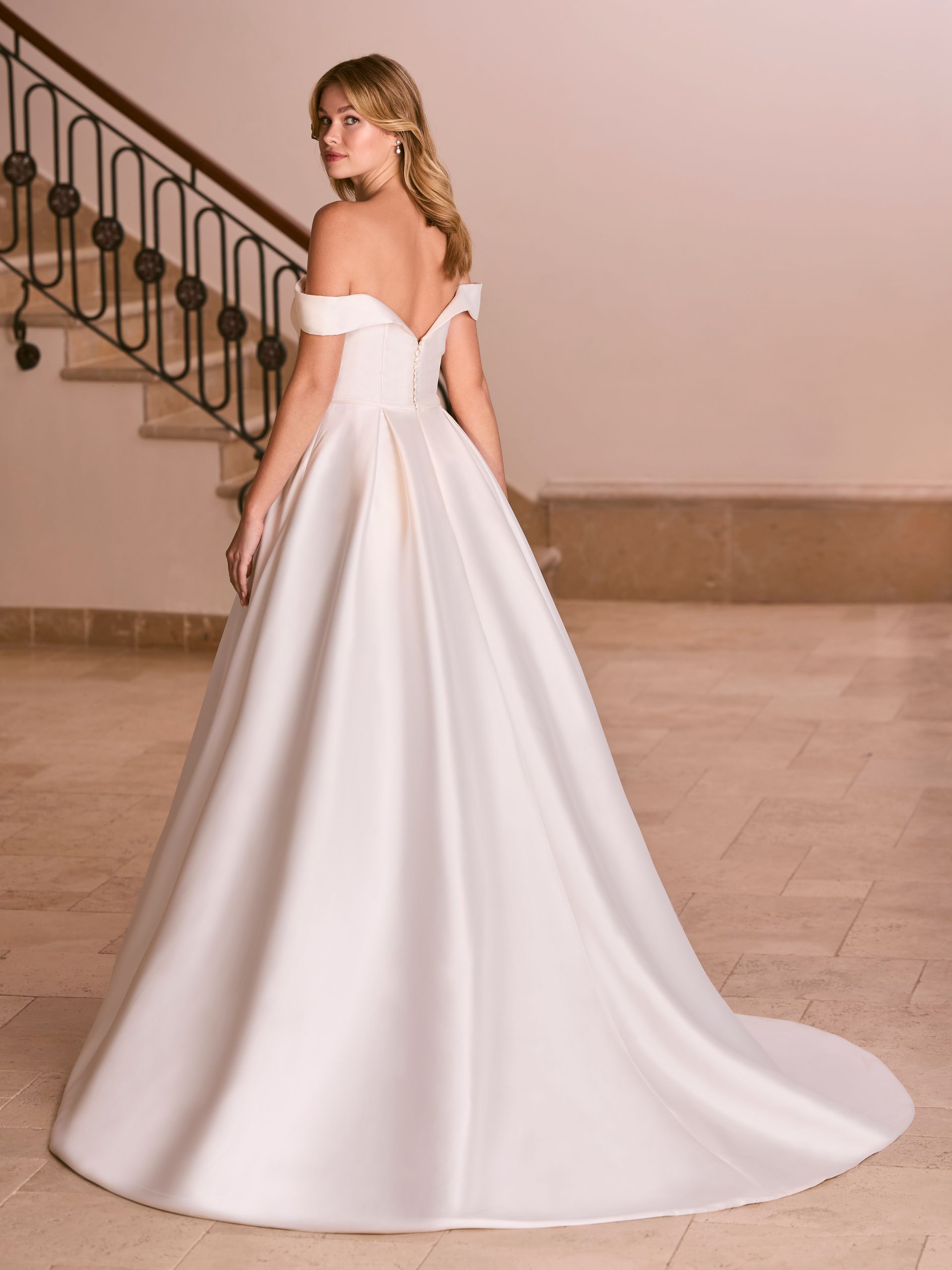 Donna in abito da sposa in raso bianco con spalle scoperte, girata con lo sguardo oltre la spalla. Sullo sfondo, la grande scalinata.