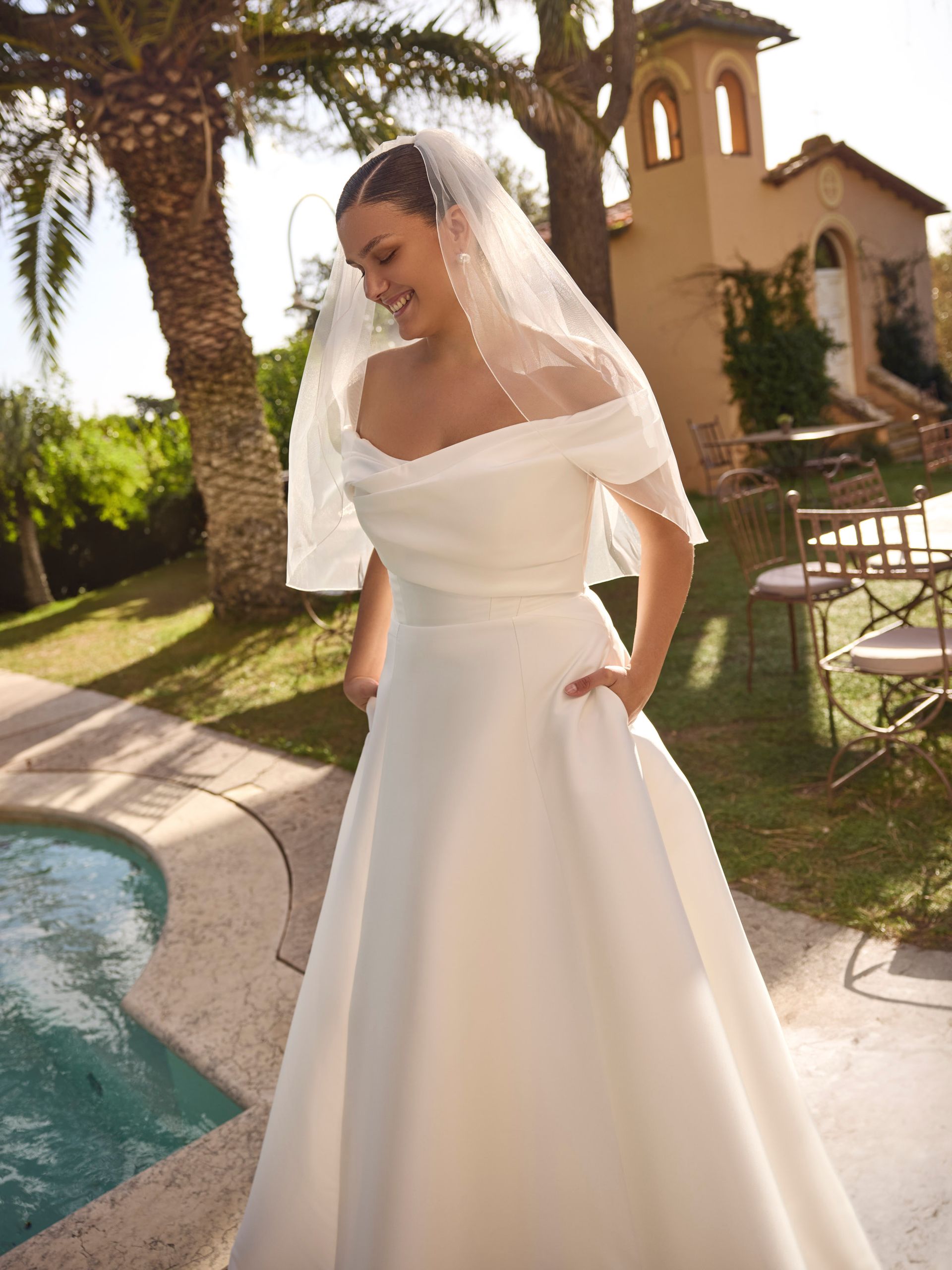 Sposa in abito bianco con velo e spalle scoperte, mani in tasca, sorridente all'aperto vicino a una piscina.