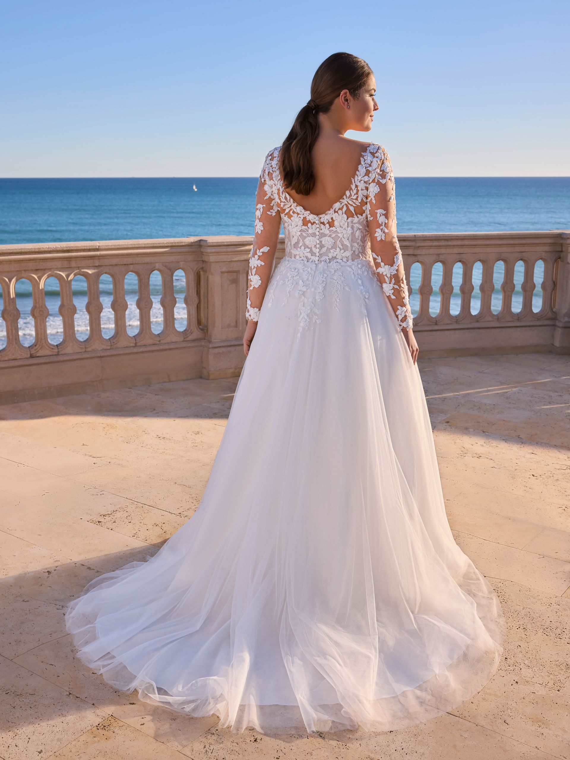 Donna in abito da sposa bianco con maniche lunghe, rivolta verso l'oceano.
