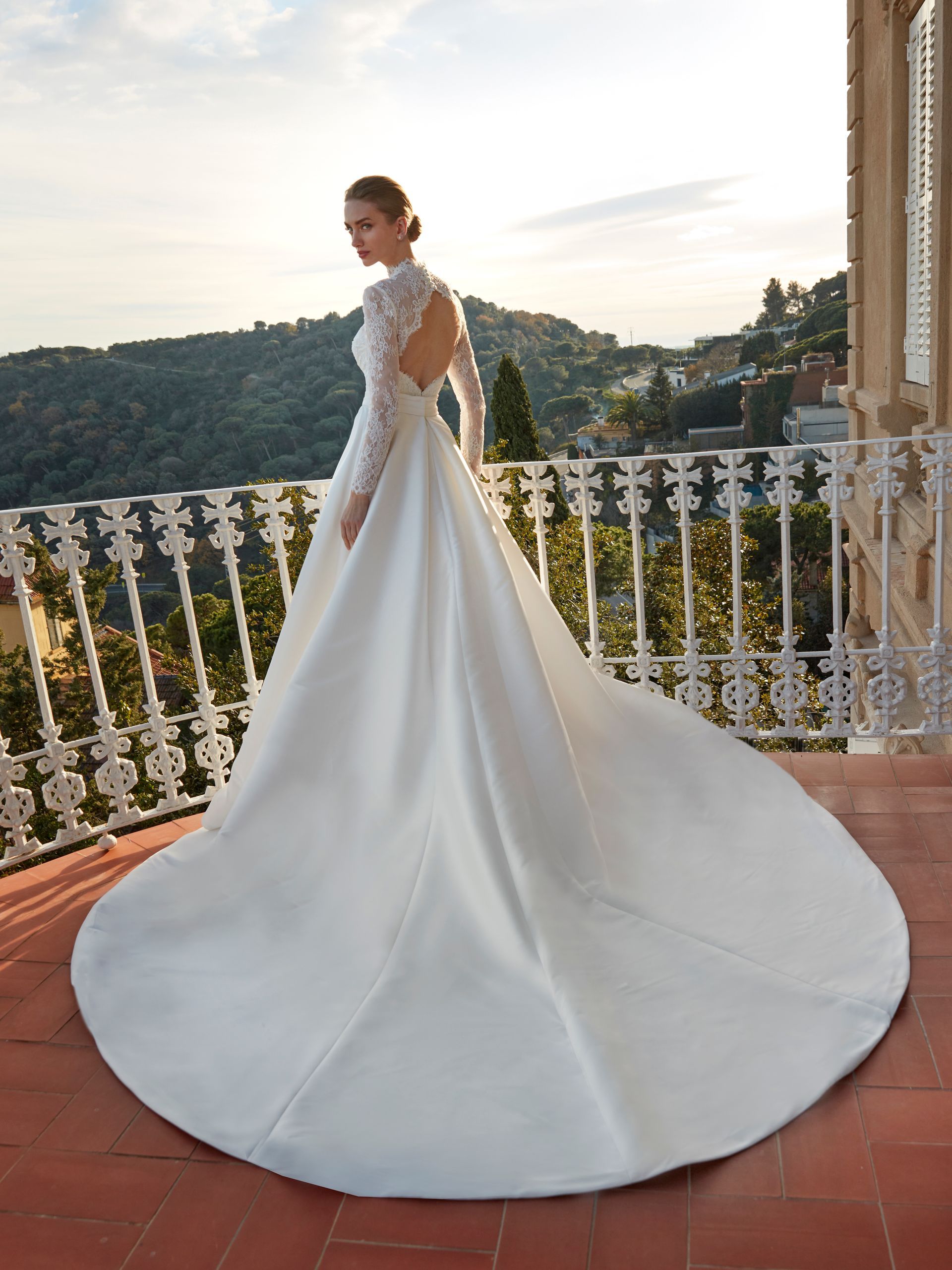 Donna in abito da sposa bianco con lungo strascico su un balcone con vista sulla città.