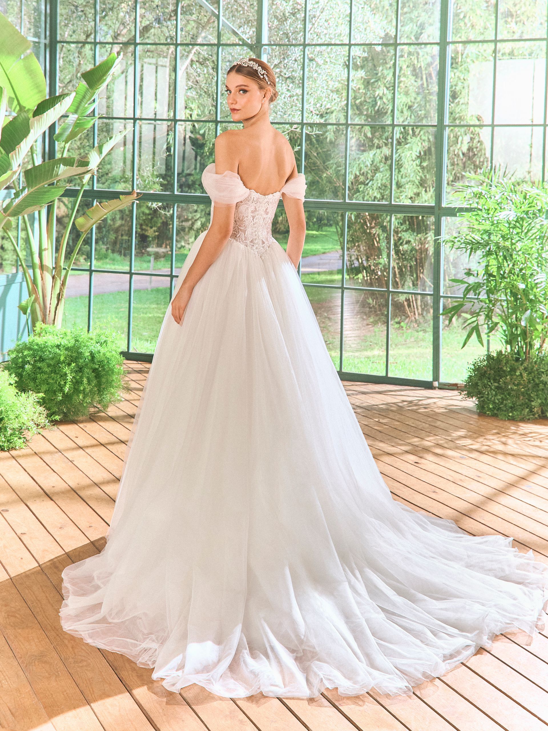 Donna in un abito da sposa bianco con spalle scoperte e lungo strascico, in piedi in una serra.