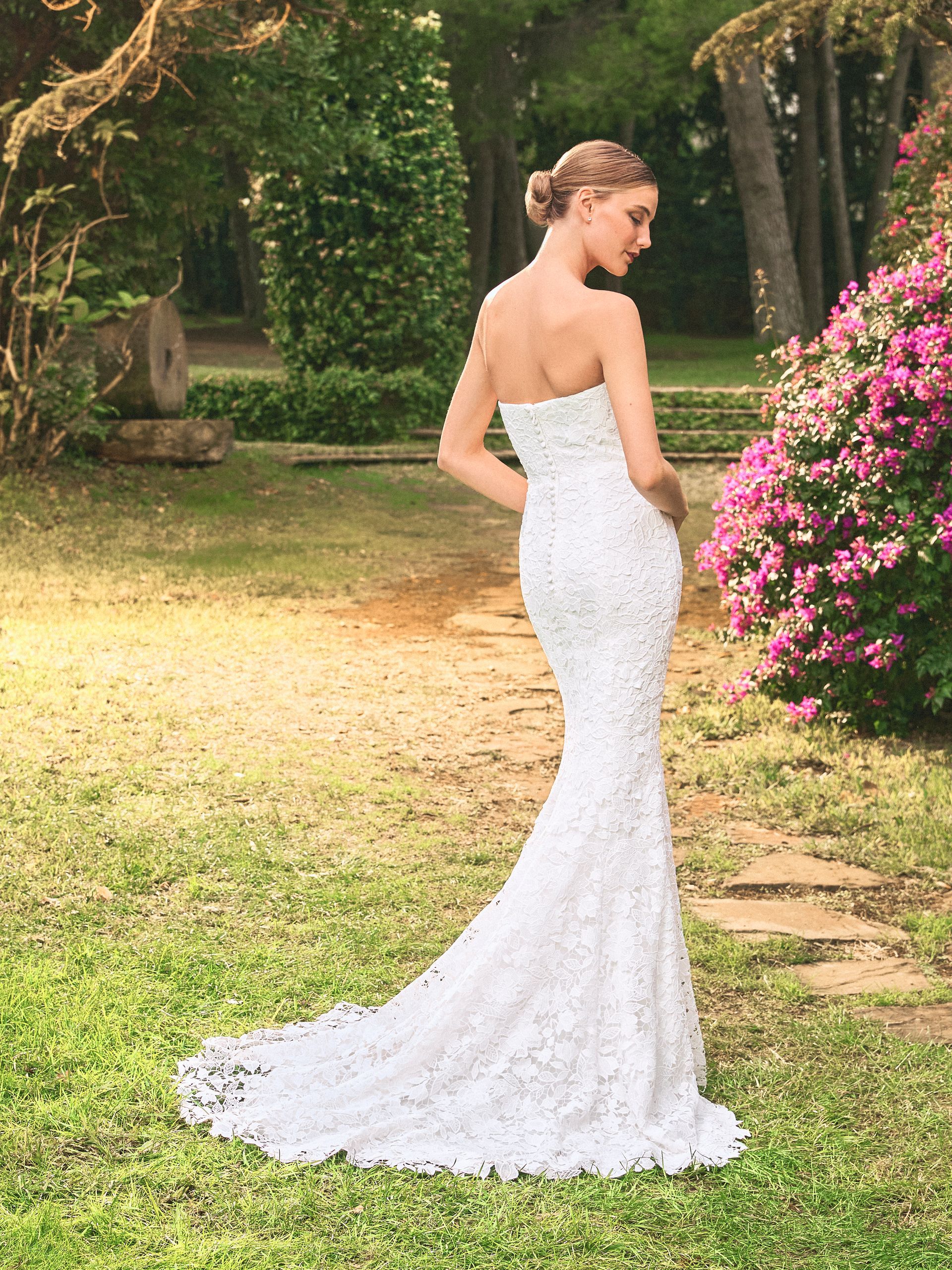 Una donna in abito da sposa bianco senza spalline in pizzo è in piedi in giardino, con lo sguardo rivolto oltre la spalla, alla luce del sole.