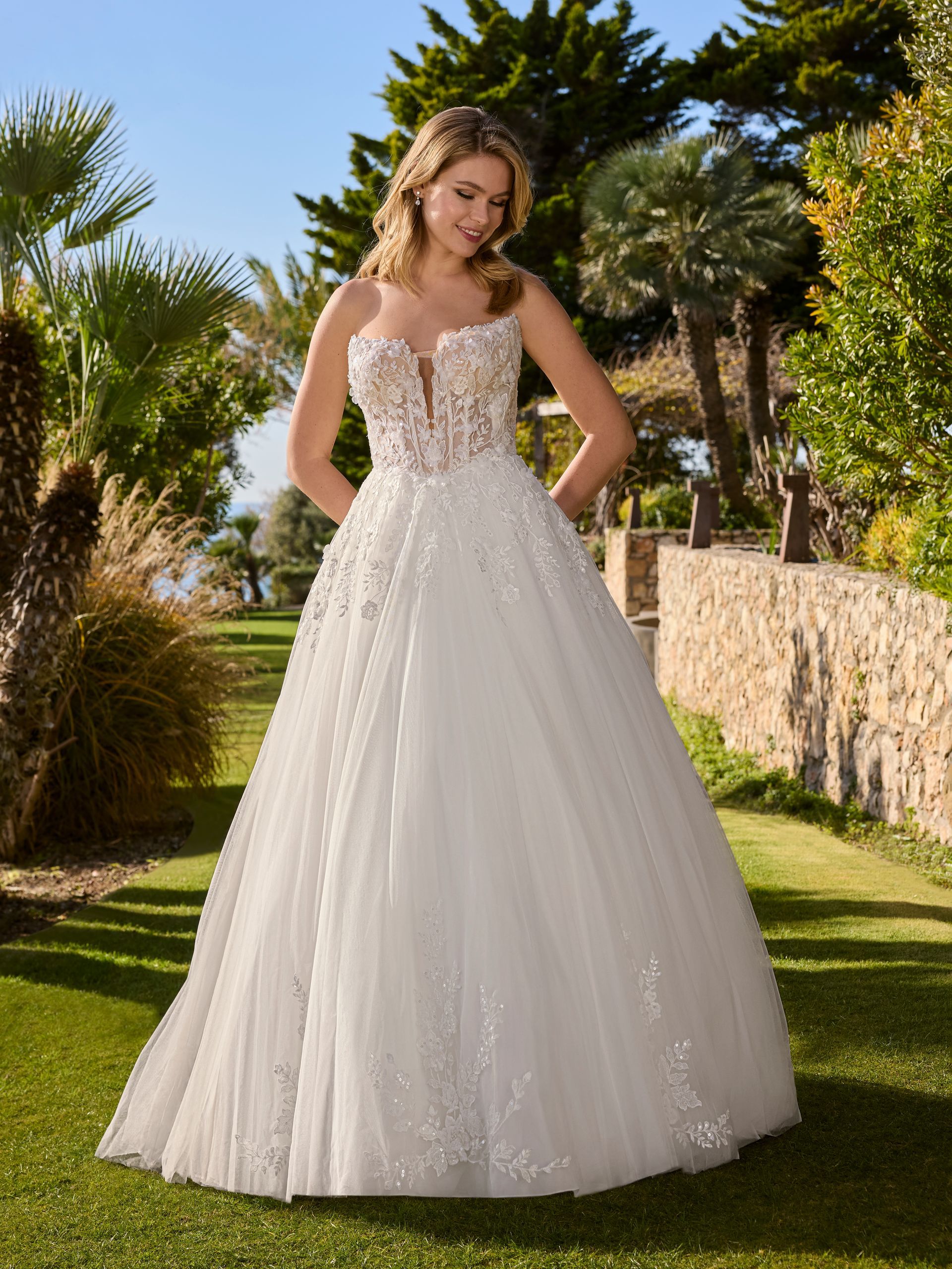 Donna in abito da sposa bianco, in piedi all'aperto con un giardino sullo sfondo, sorridente.