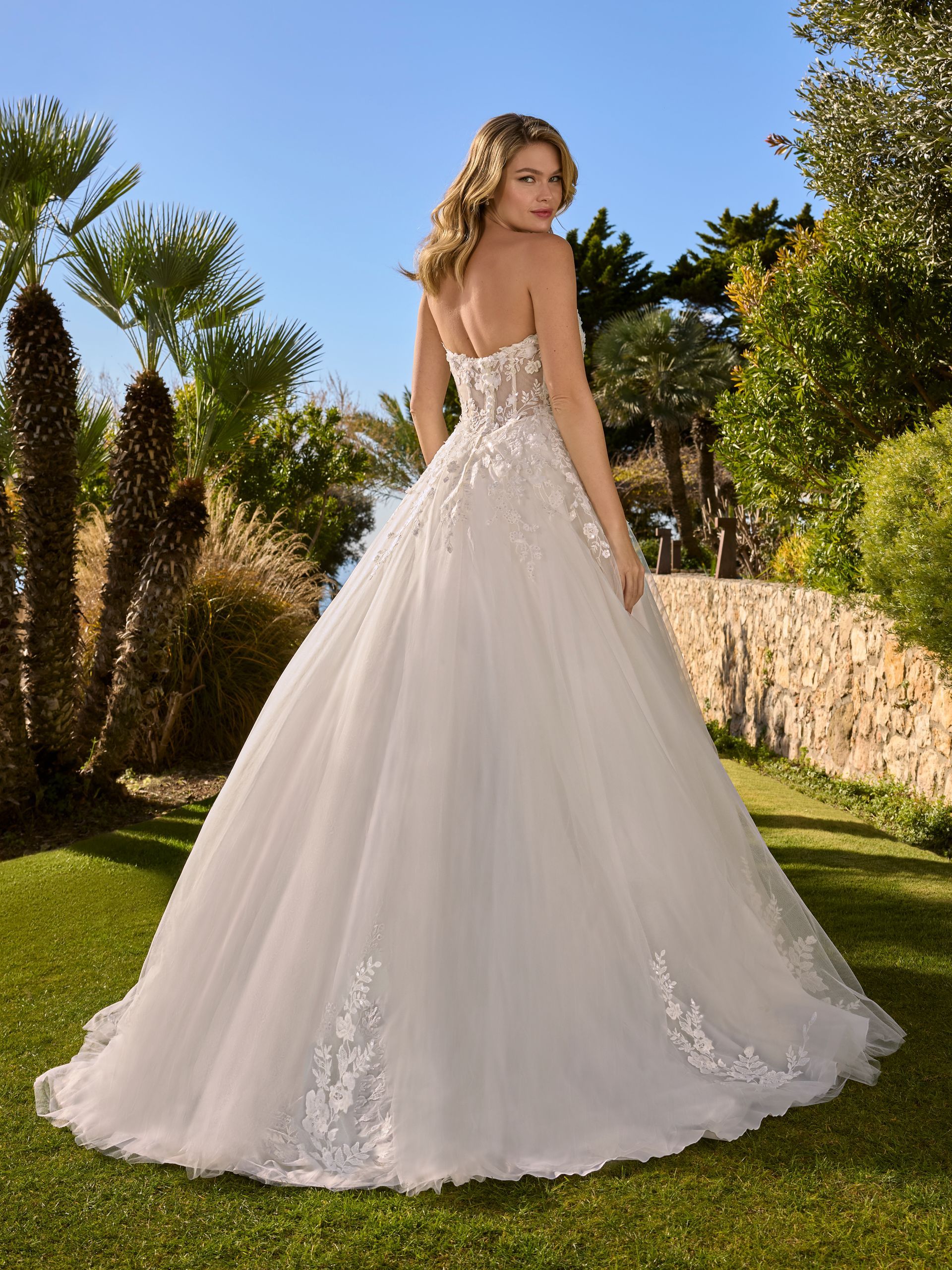 Una donna in abito da sposa bianco senza spalline posa all'aperto, di spalle alla telecamera, con giardino e cielo azzurro sullo sfondo.