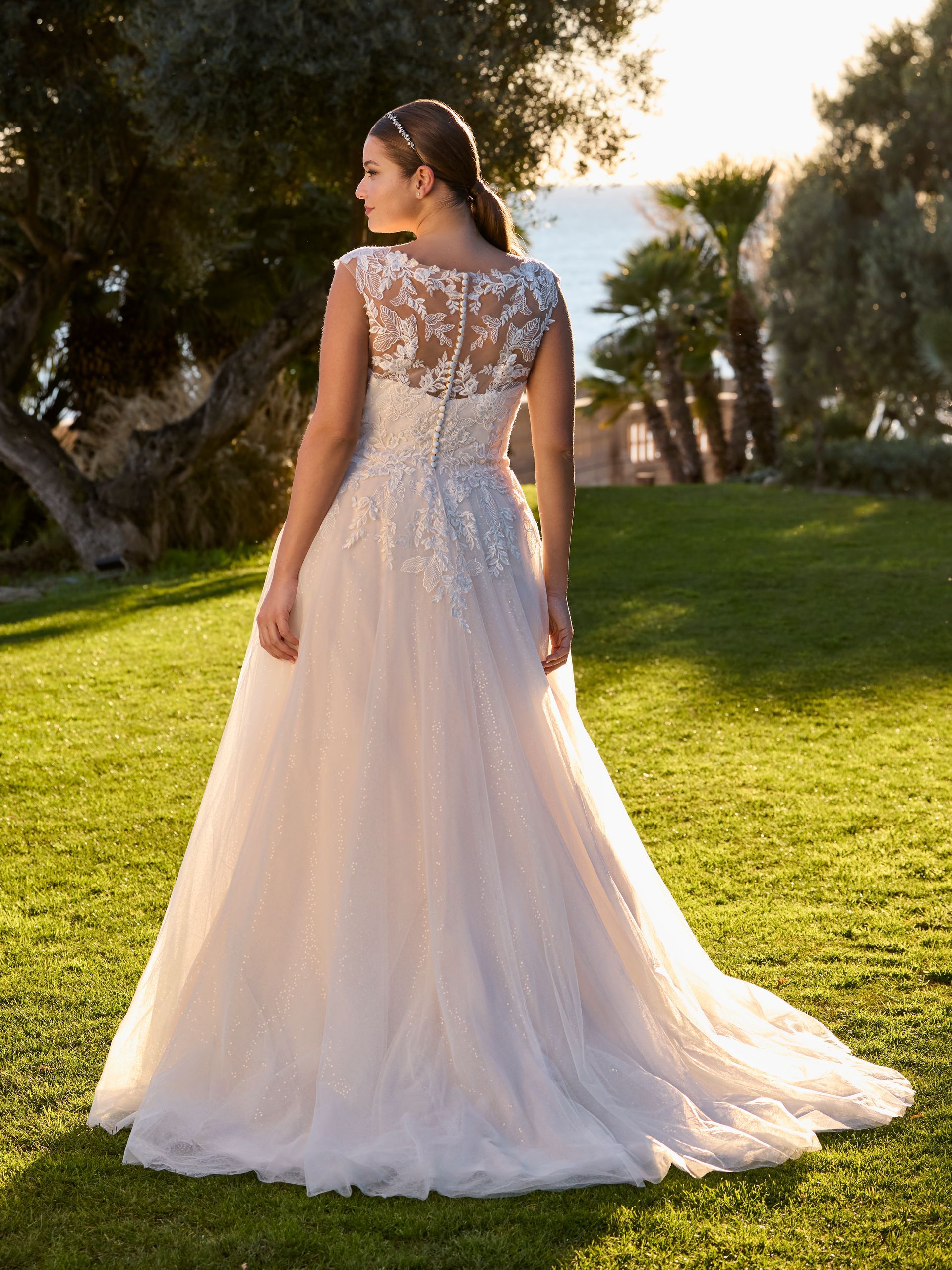 Donna in abito da sposa bianco all'aperto, spalle alla telecamera, corpetto in pizzo, lungo strascico, giornata di sole.