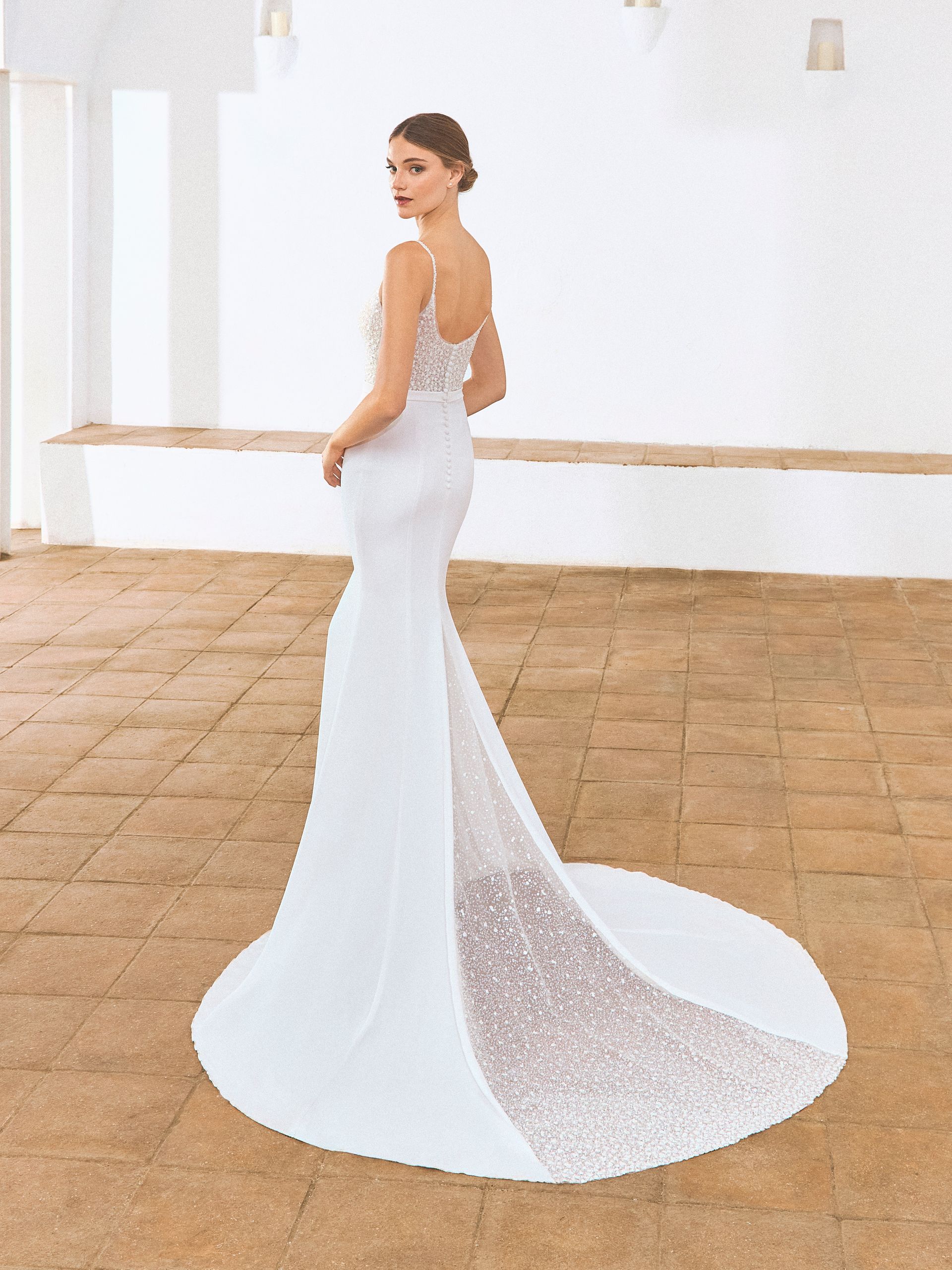 Donna in abito da sposa bianco con lungo strascico, in piedi in una stanza piena di luce, che guarda oltre la sua spalla.