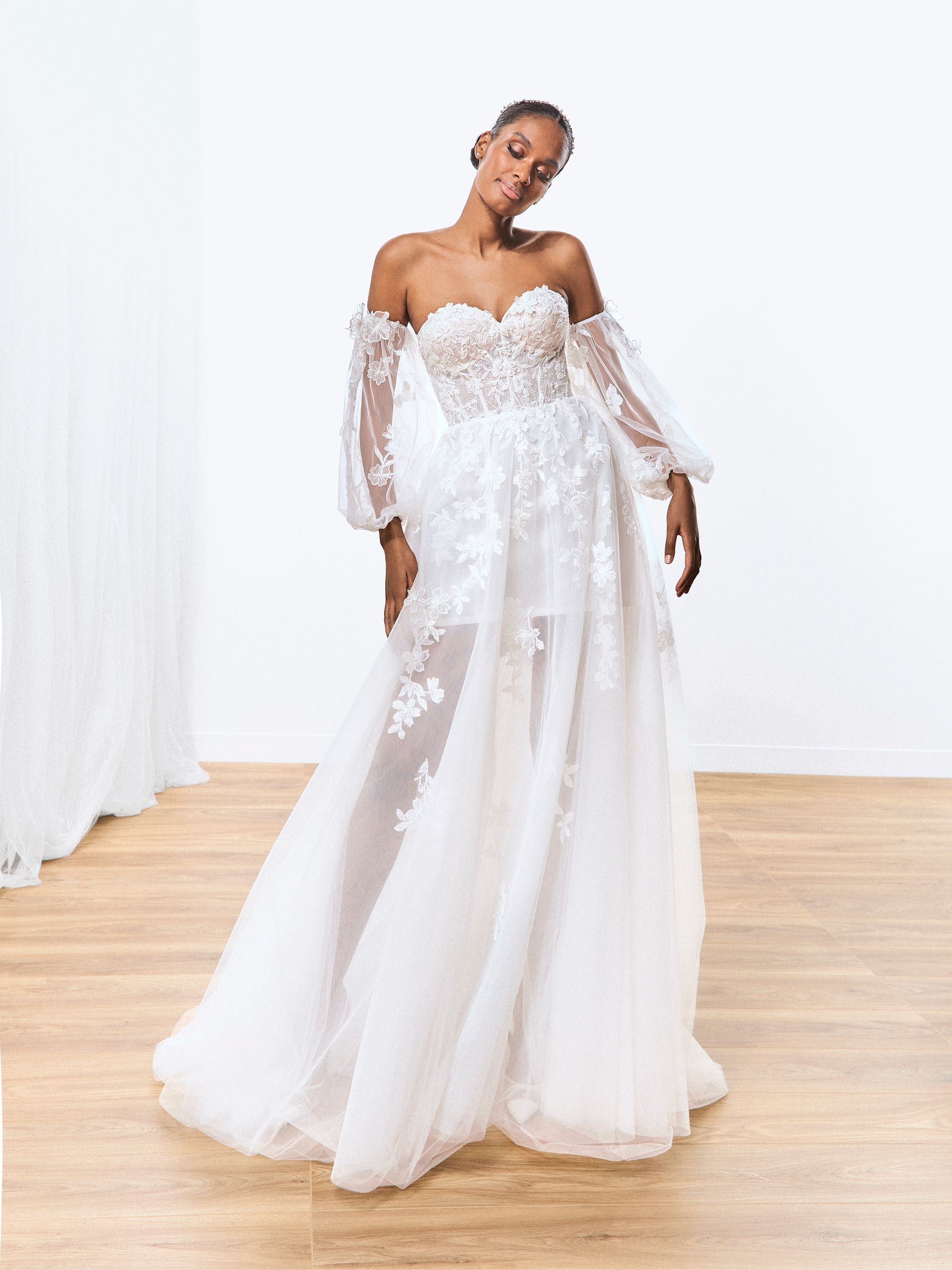 Donna in abito da sposa bianco con spalle scoperte e decorazioni floreali. In piedi in uno studio con pavimento in legno chiaro.