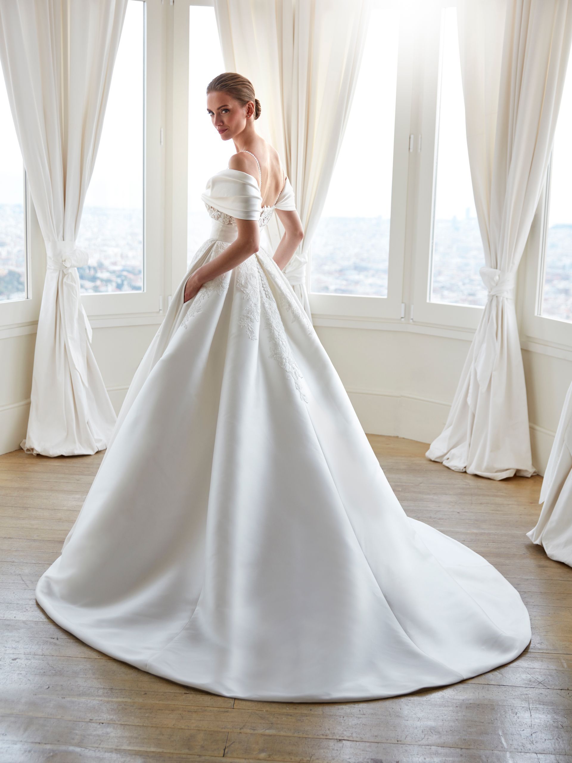 Donna con un abito da sposa bianco con le spalle scoperte, in piedi in una stanza luminosa con tende drappeggiate.