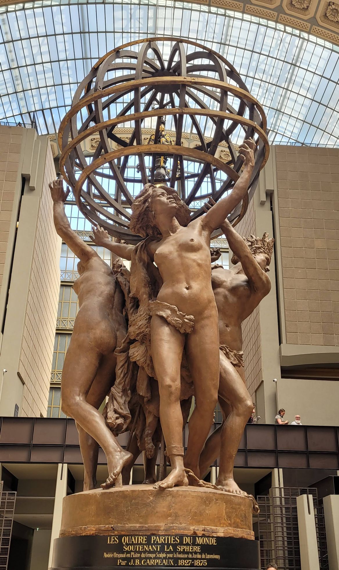 Statua in bronzo raffigurante figure nude che sorreggono un grande globo. Situata all'interno di un edificio con soffitto in vetro.