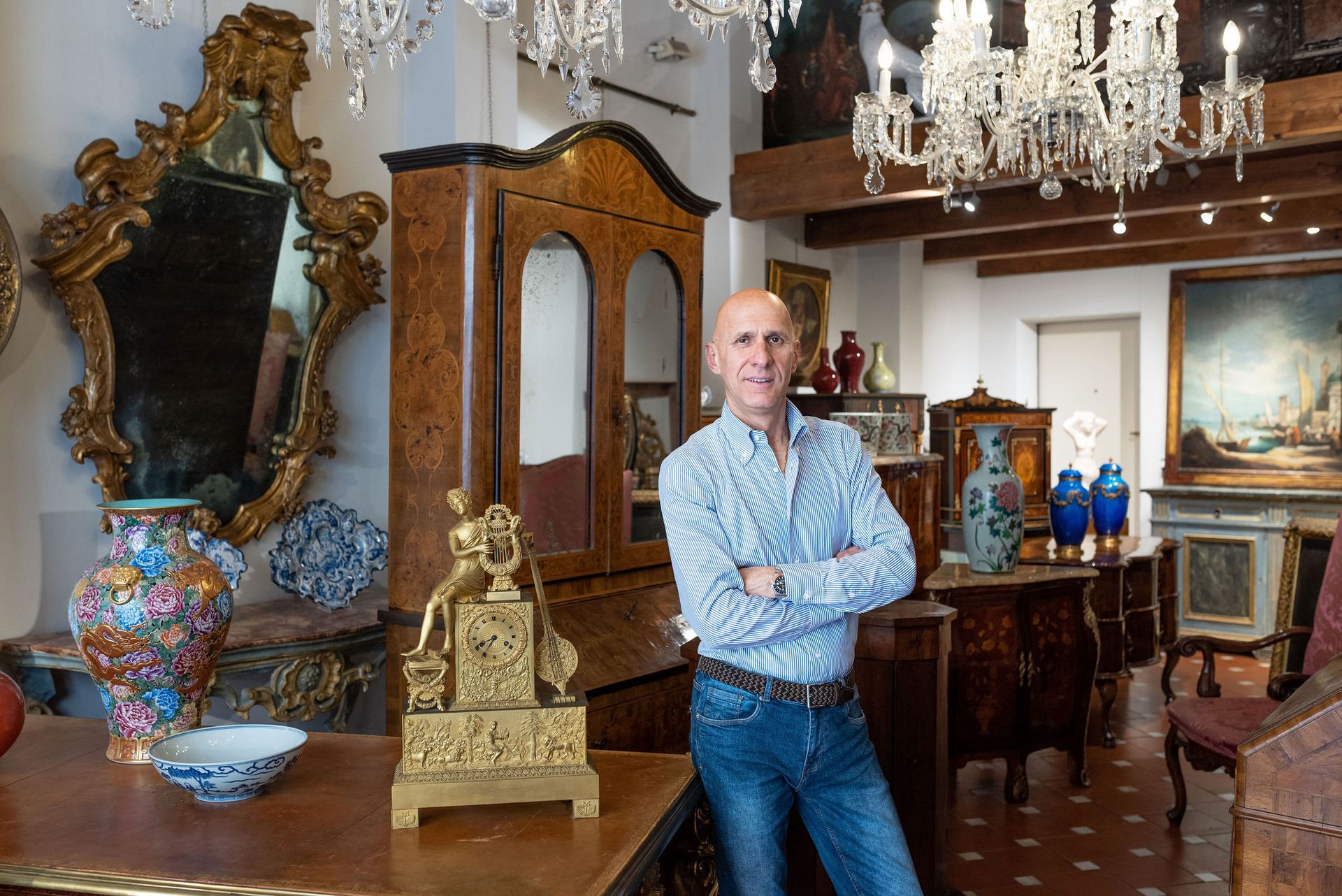 Uomo in un negozio di antiquariato, braccia incrociate, circondato da mobili e lampadari.