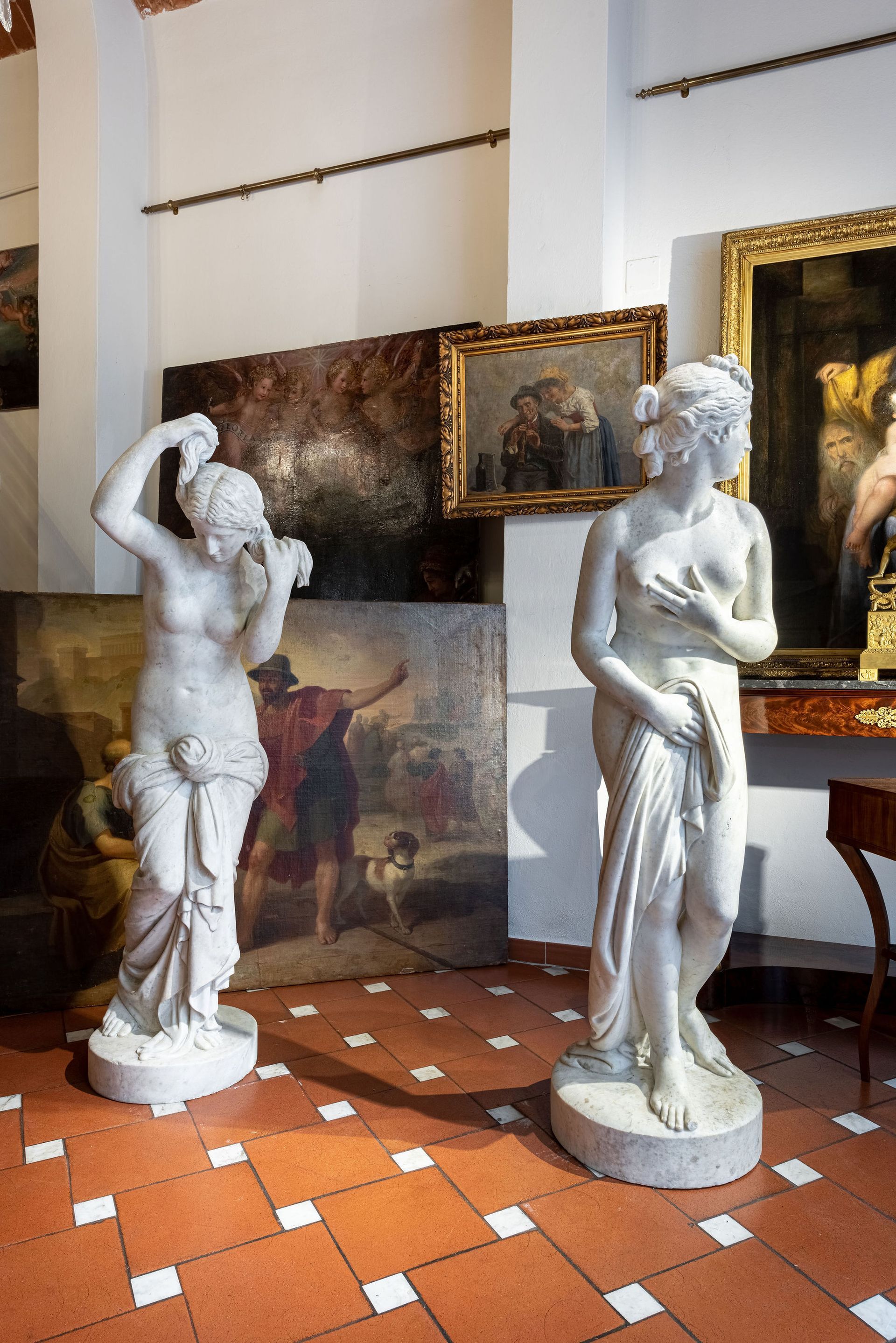 In una stanza, su un pavimento piastrellato, si trovano due sculture di donne in marmo bianco, con dei dipinti sullo sfondo.