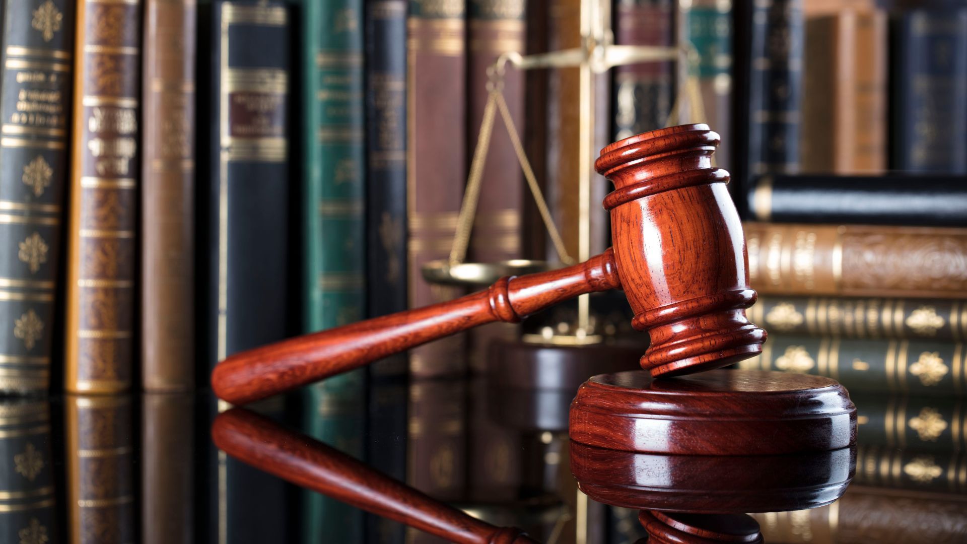 Wooden gavel on a sound block, with a scale and law books in the background, reflecting on a dark surface.