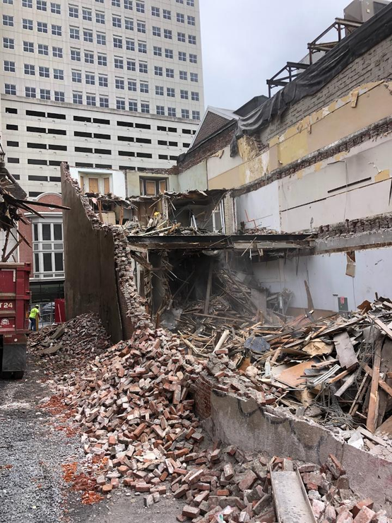 A building is being demolished in a city with a large building in the background.