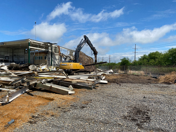 A large yellow excavator is demolishing a building in a gravel lot.